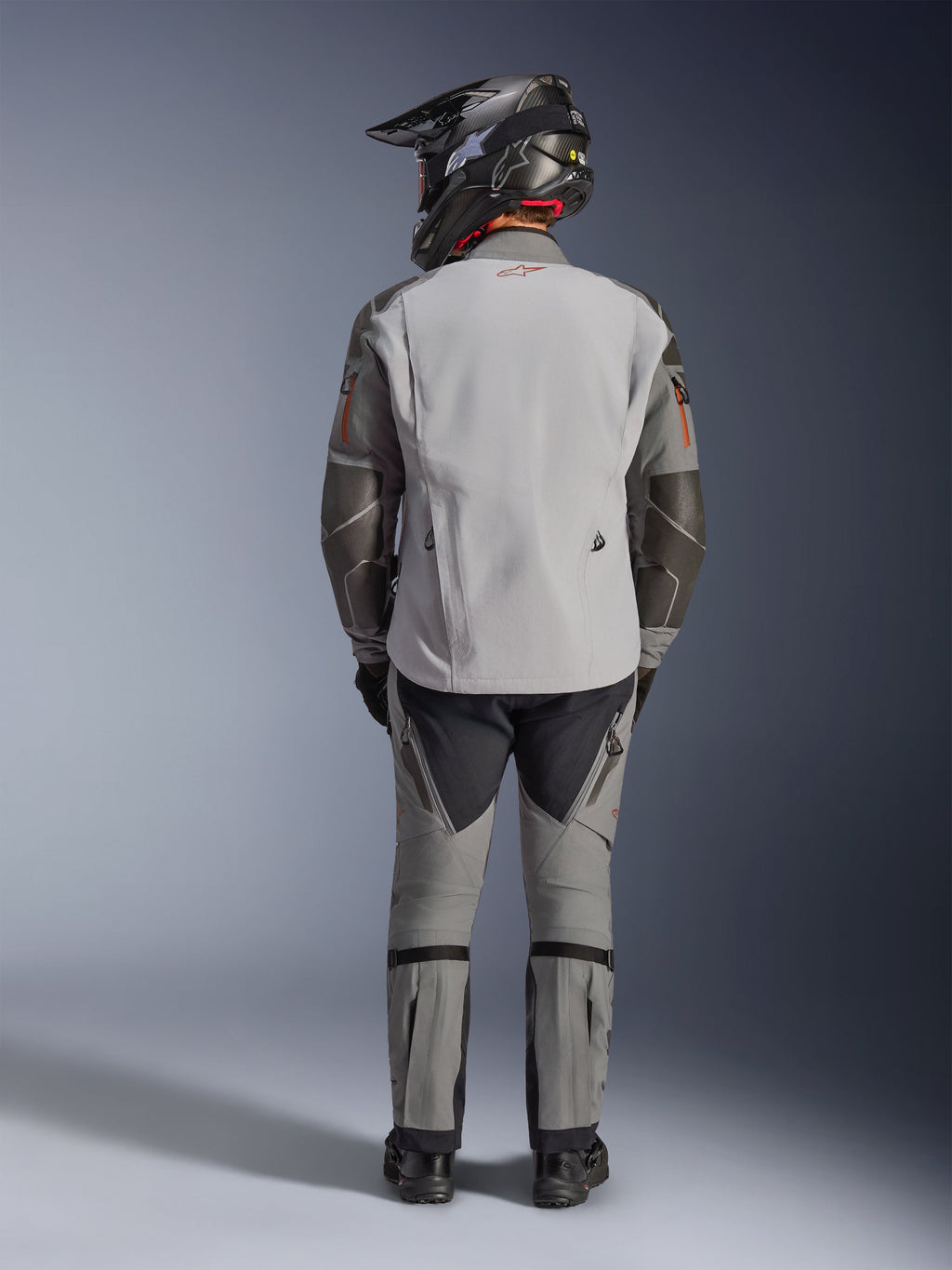 Chaqueta Alpinestars Nazca 3L GORE-TEX PRO, chaqueta de moto de aventura, gris oscuro y gris medio, vista trasera del piloto con equipamiento completo, combinada con pantalones a juego y casco de aventura negro, con logo rojo en la espalda y refuerzos de Superfabric en los codos, diseño textil duradero para turismo de aventura.