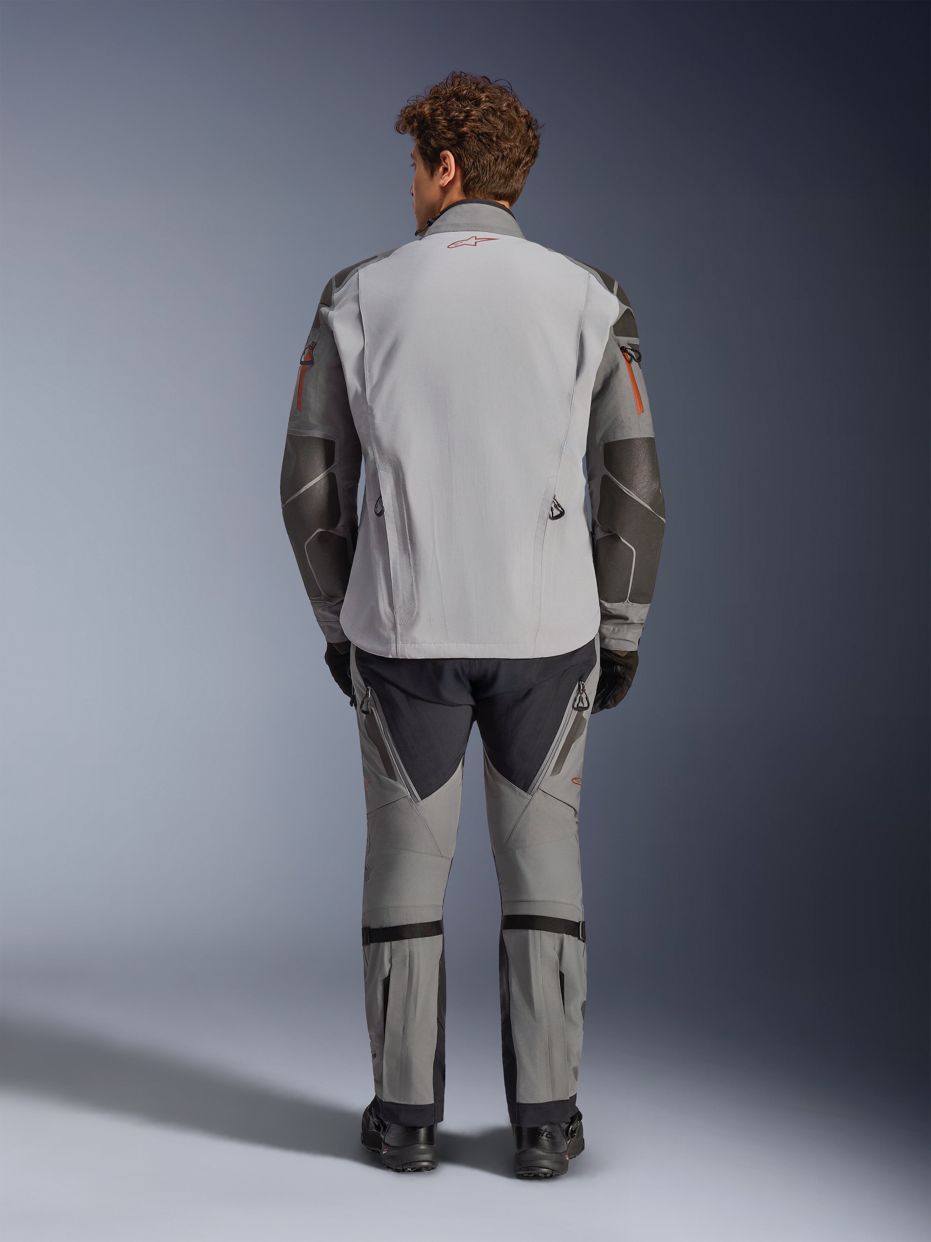 Chaqueta Alpinestars Nazca 3L GORE-TEX PRO, vista trasera, gris oscuro y gris medio, chaqueta de moto de aventura con construcción GORE-TEX, logo rojo en el cuello, detalles naranjas en las mangas, usada con pantalones de conducción a juego y guantes negros.