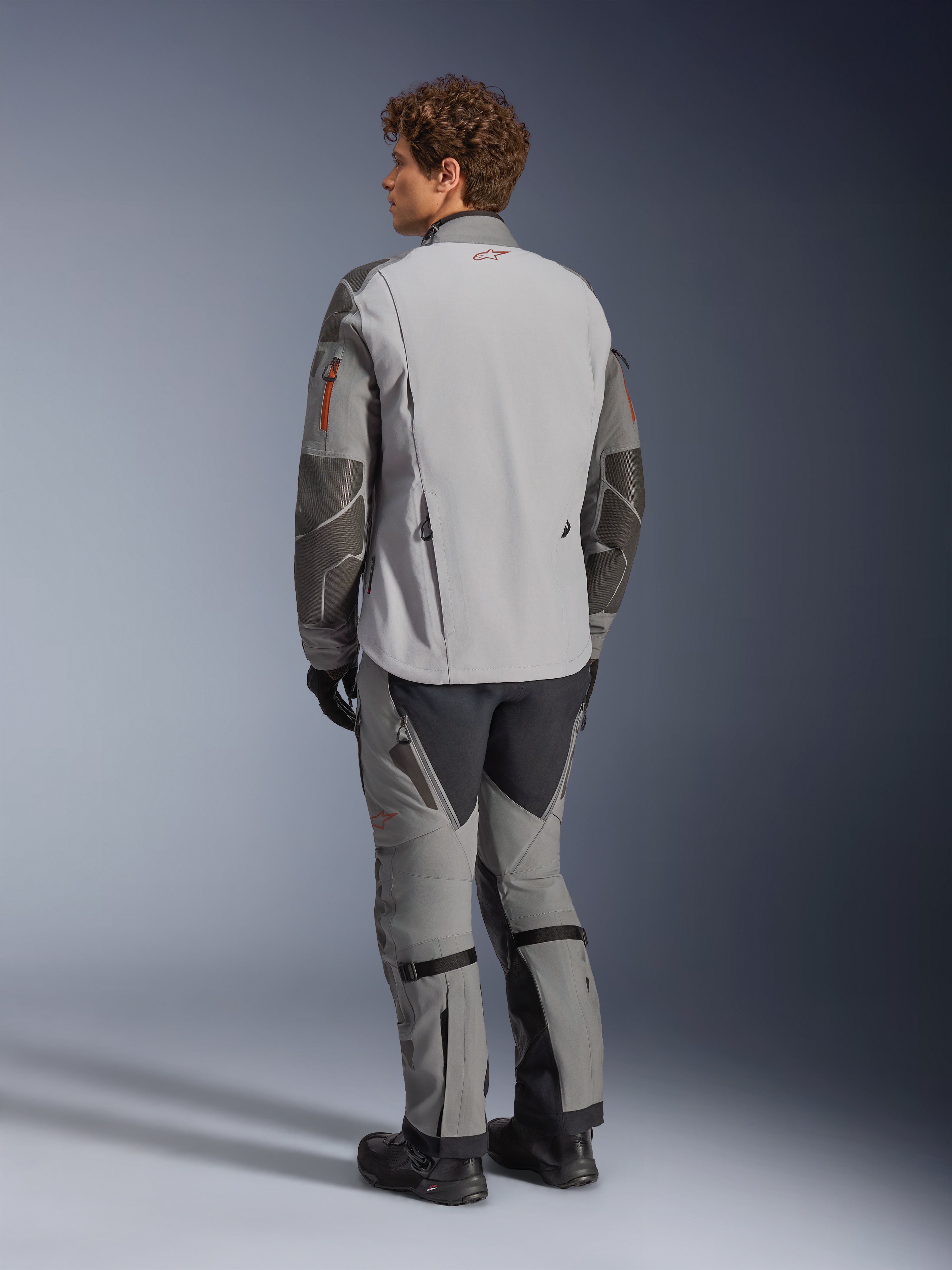 Chaqueta Alpinestars Nazca 3L GORE-TEX PRO, chaqueta de moto de aventura, en gris oscuro y gris medio, vista trasera, usada con pantalones de aventura a juego y botas negras, con refuerzos de Superfabric gris oscuro en las mangas, detalles de cremalleras en naranja y un pequeño logo rojo en la parte posterior del cuello.