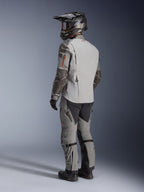 Chaqueta Alpinestars Nazca 3L GORE-TEX PRO, chaqueta de moto de aventura en gris oscuro y gris medio, vista trasera de tres cuartos en modelo con equipamiento completo, incluyendo pantalones y casco a juego, diseño técnico con refuerzos Superfabric en los codos.