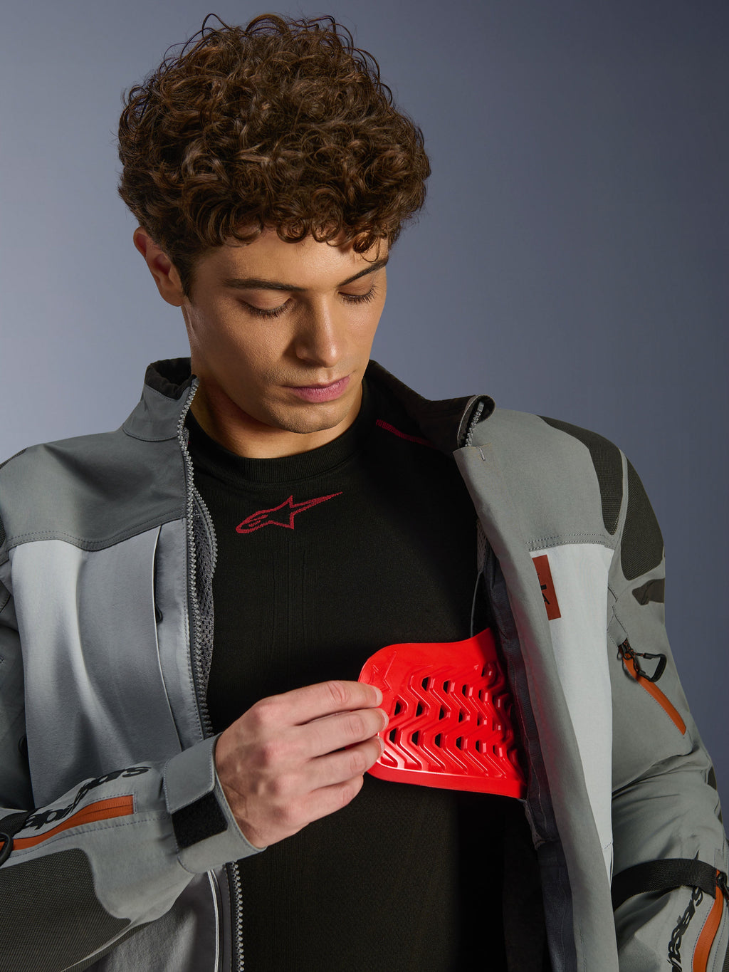 Modelo masculino luciendo la chaqueta Alpinestars Nazca 3L GORE-TEX PRO en gris oscuro y gris medio, mostrando el inserto de protección rojo Nucleon PLASMA Pro, ropa de conducción de aventura, chaqueta de moto premium con detalles técnicos