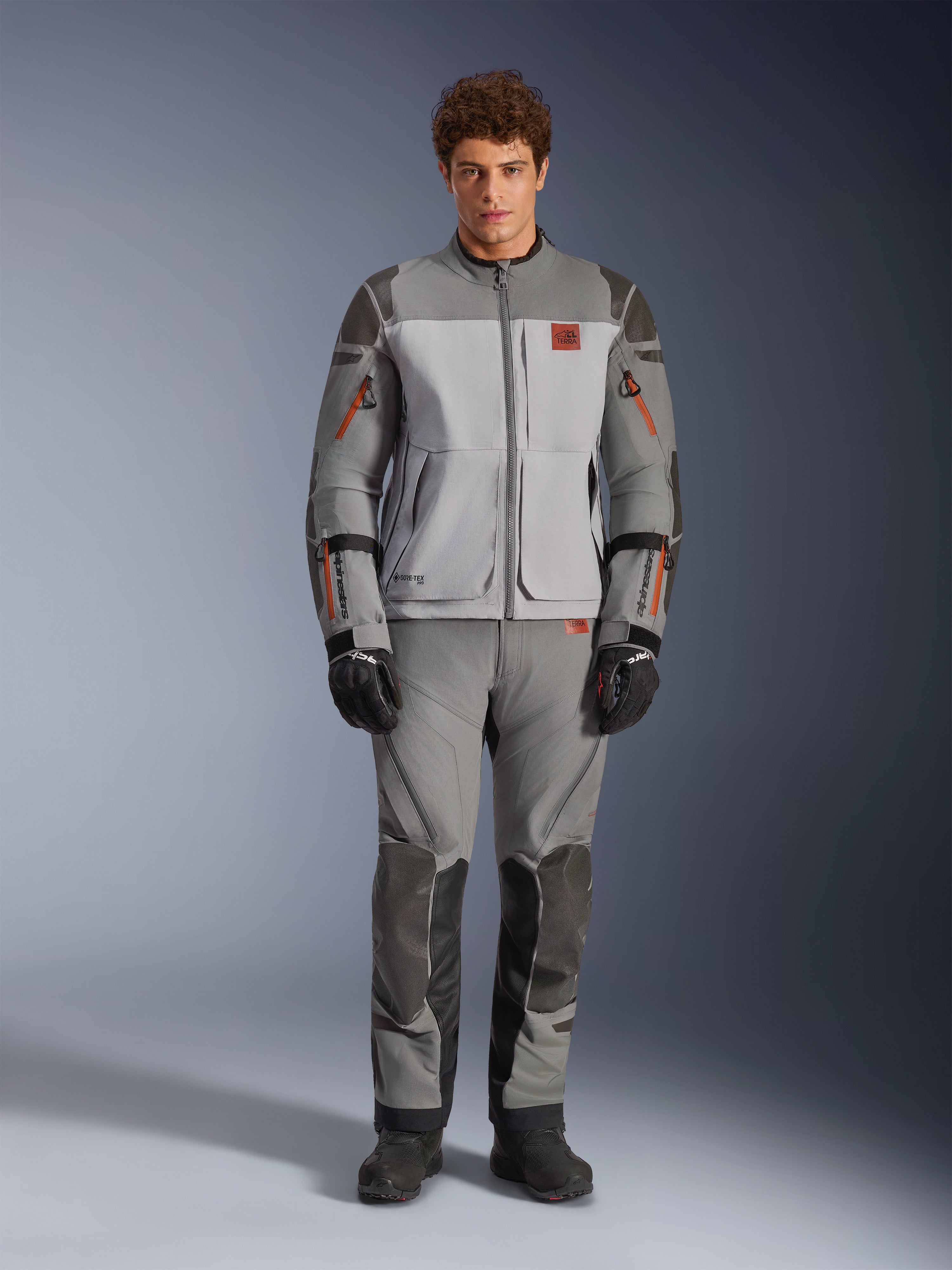 Chaqueta Alpinestars Nazca 3L GORE-TEX PRO, chaqueta de moto de aventura, gris oscuro y gris medio, vestida por un modelo con pantalones a juego y guantes negros, con detalles de cremalleras en naranja y logotipo rojo en el pecho, equipo de protección técnica para conducción extrema
