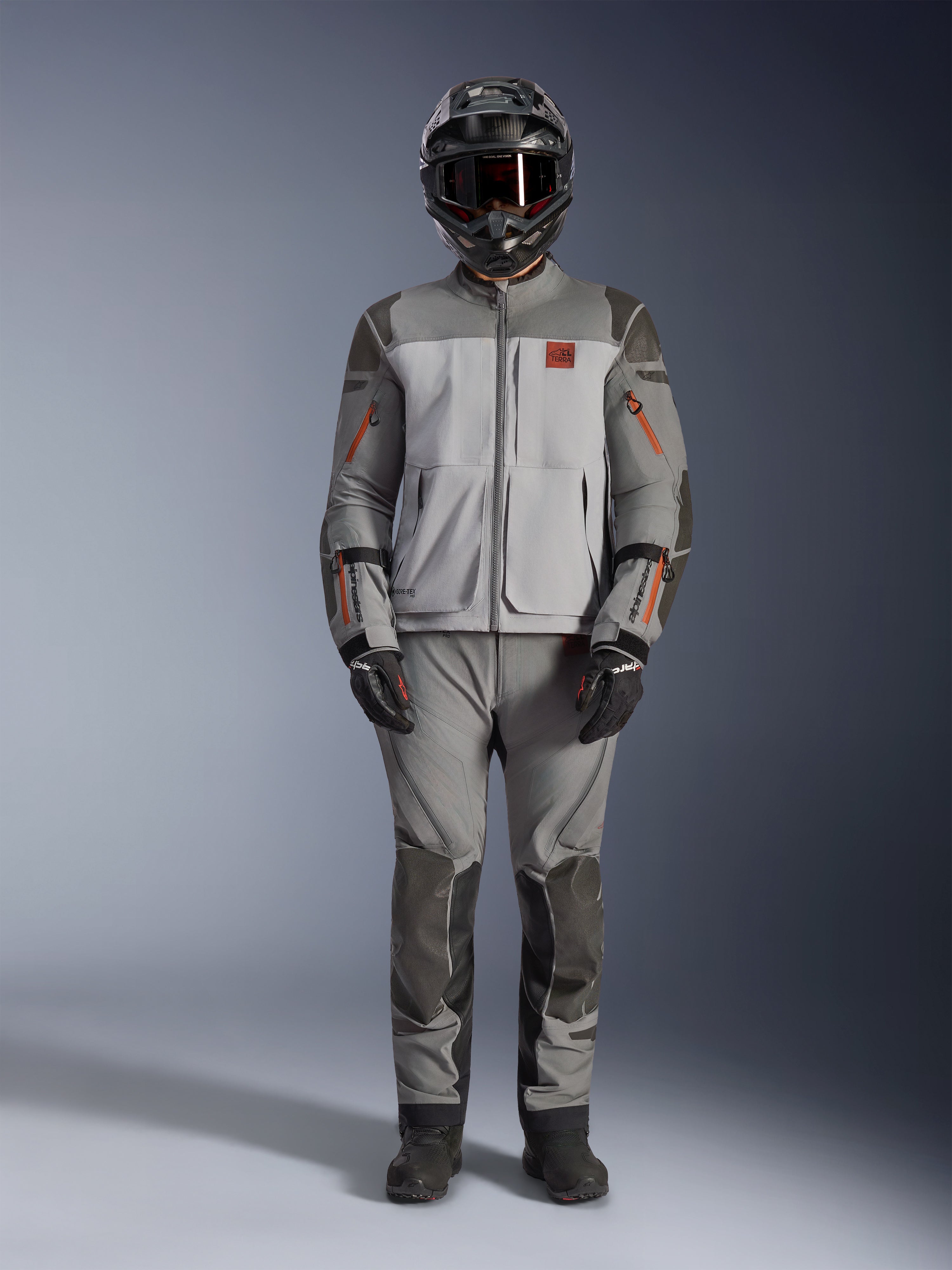 Chaqueta Alpinestars Nazca 3L GORE-TEX PRO, chaqueta de moto de aventura, gris oscuro y gris medio con detalles en naranja, con refuerzos de Superfabric y cremalleras de ventilación frontal, usada con pantalones a juego y casco negro, equipamiento técnico para conducción todo terreno