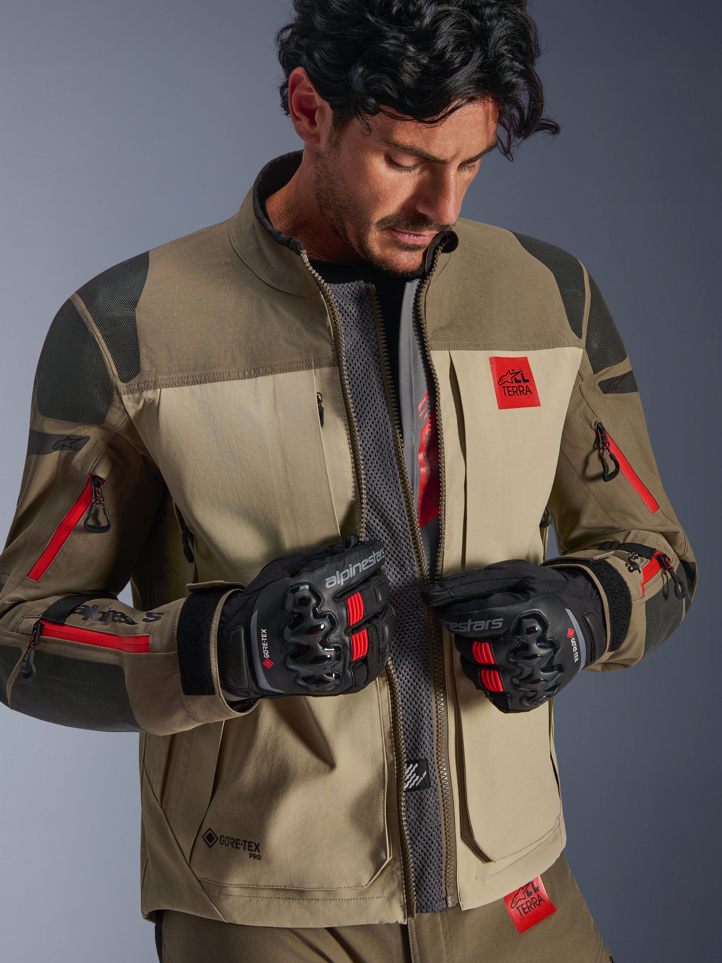 Chaqueta Alpinestars Nazca 3L GORE-TEX PRO, Walnut Rock Brown, chaqueta de moto de aventura con el frente abierto revelando el panel central de malla para el flujo de aire, logotipo All Terra en rojo, usada con guantes de moto GORE-TEX negros, equipamiento técnico para touring off-road
