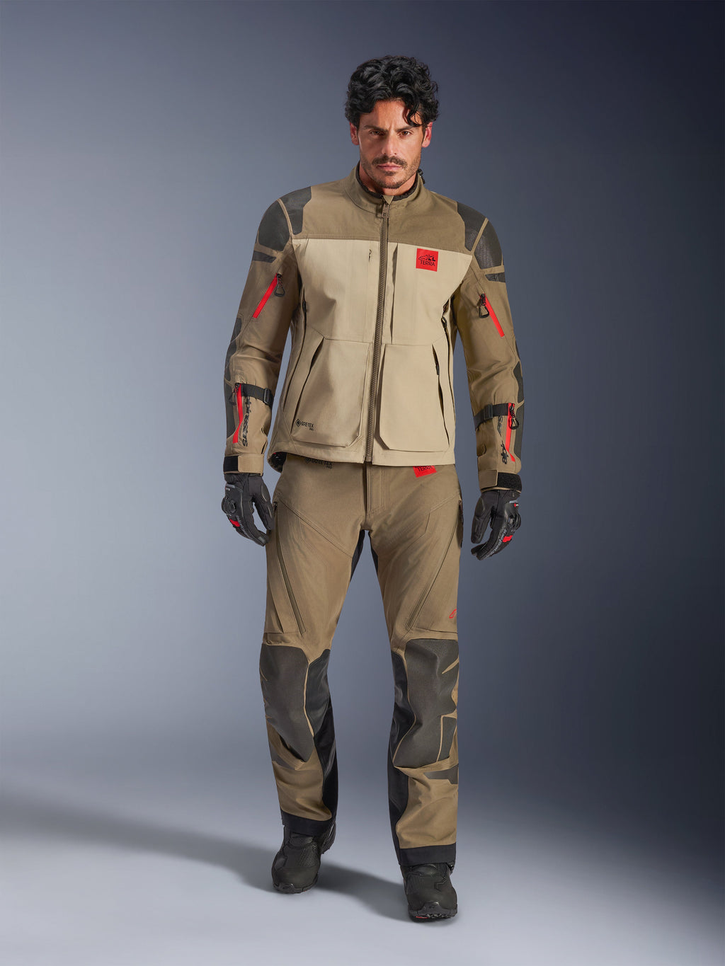 Chaqueta Alpinestars Nazca 3L GORE-TEX PRO, Walnut Rock Brown, chaqueta de motociclismo de aventura usada con pantalones y guantes a juego, con detalles de cremalleras en rojo, logotipo 'All Terra' en el pecho y construcción textil robusta.