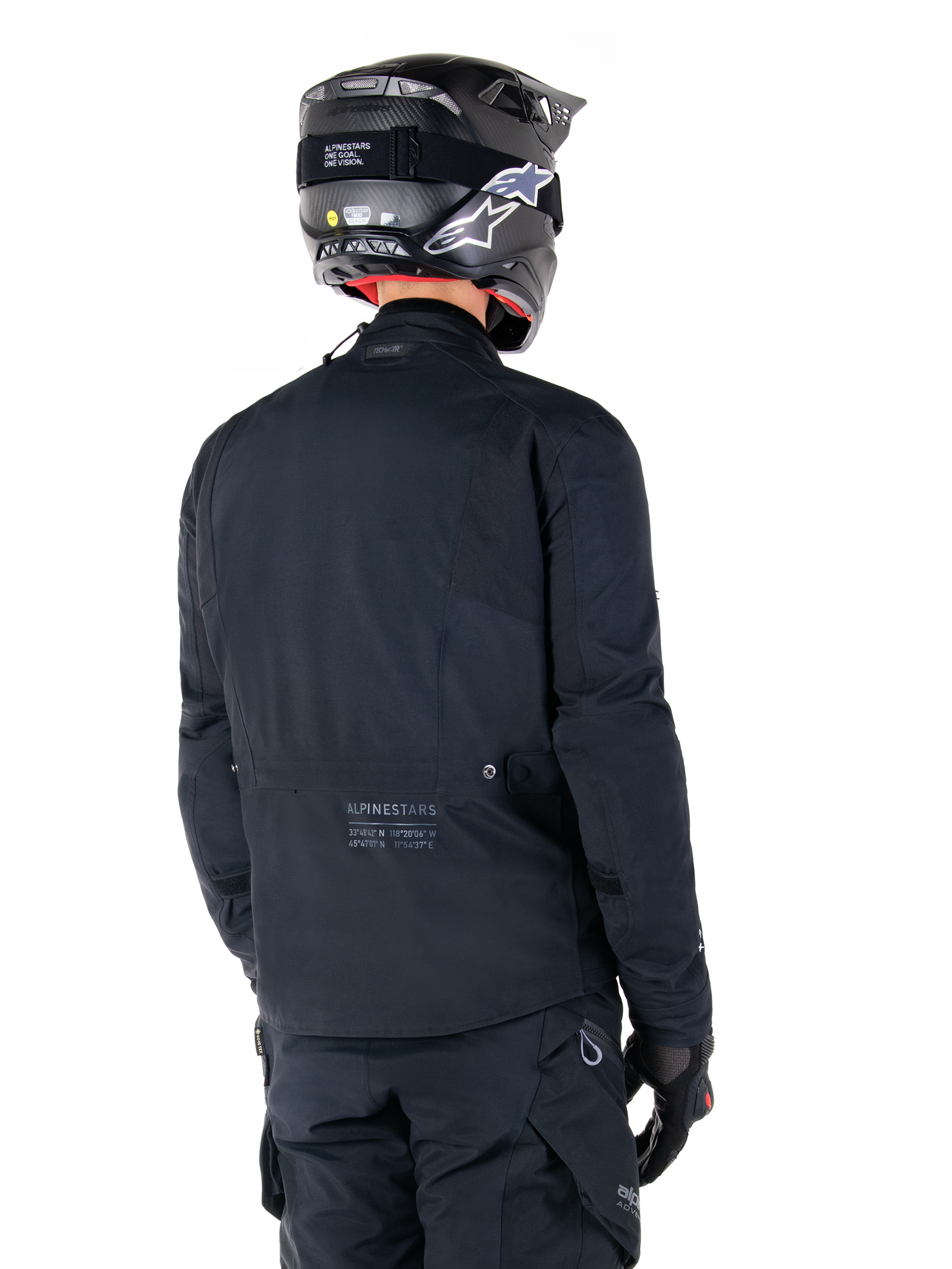 Alpinestars St-7 Prime GORE-TEX Jacket, Black, rear view of a rider wearing the jacket with a matching helmet and pants, featuring Alpinestars branding and geographic coordinates on the lower back.