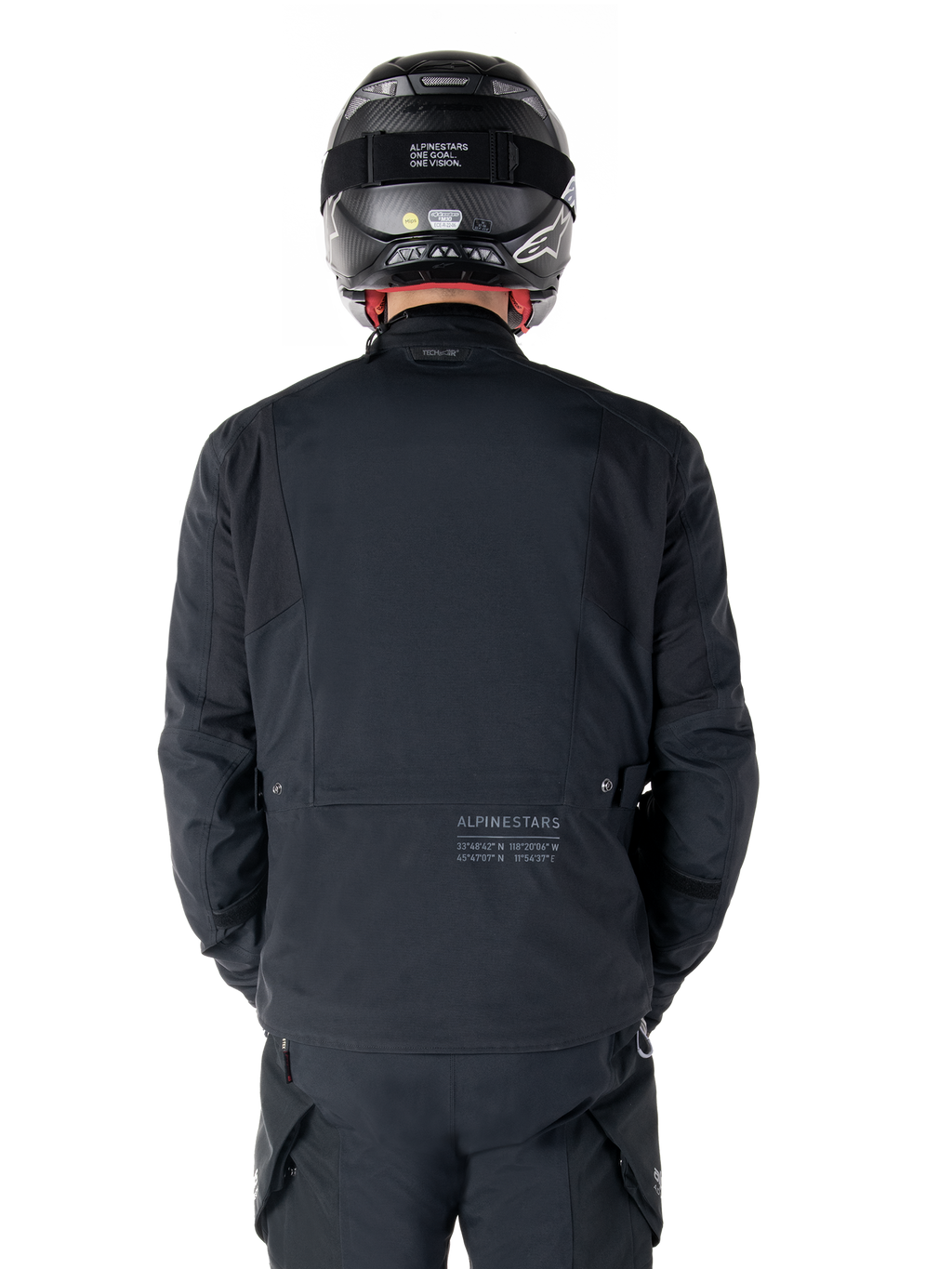 Alpinestars St-7 Prime GORE-TEX Jacket, Black, rear view of a rider, featuring subtle logo and geographic coordinates on the lower back, worn with a matching black helmet and pants, sleek and technical motorcycling apparel