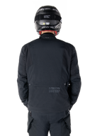 Alpinestars St-7 Prime GORE-TEX Jacket, Black, rear view of a rider, featuring subtle logo and geographic coordinates on the lower back, worn with a matching black helmet and pants, sleek and technical motorcycling apparel