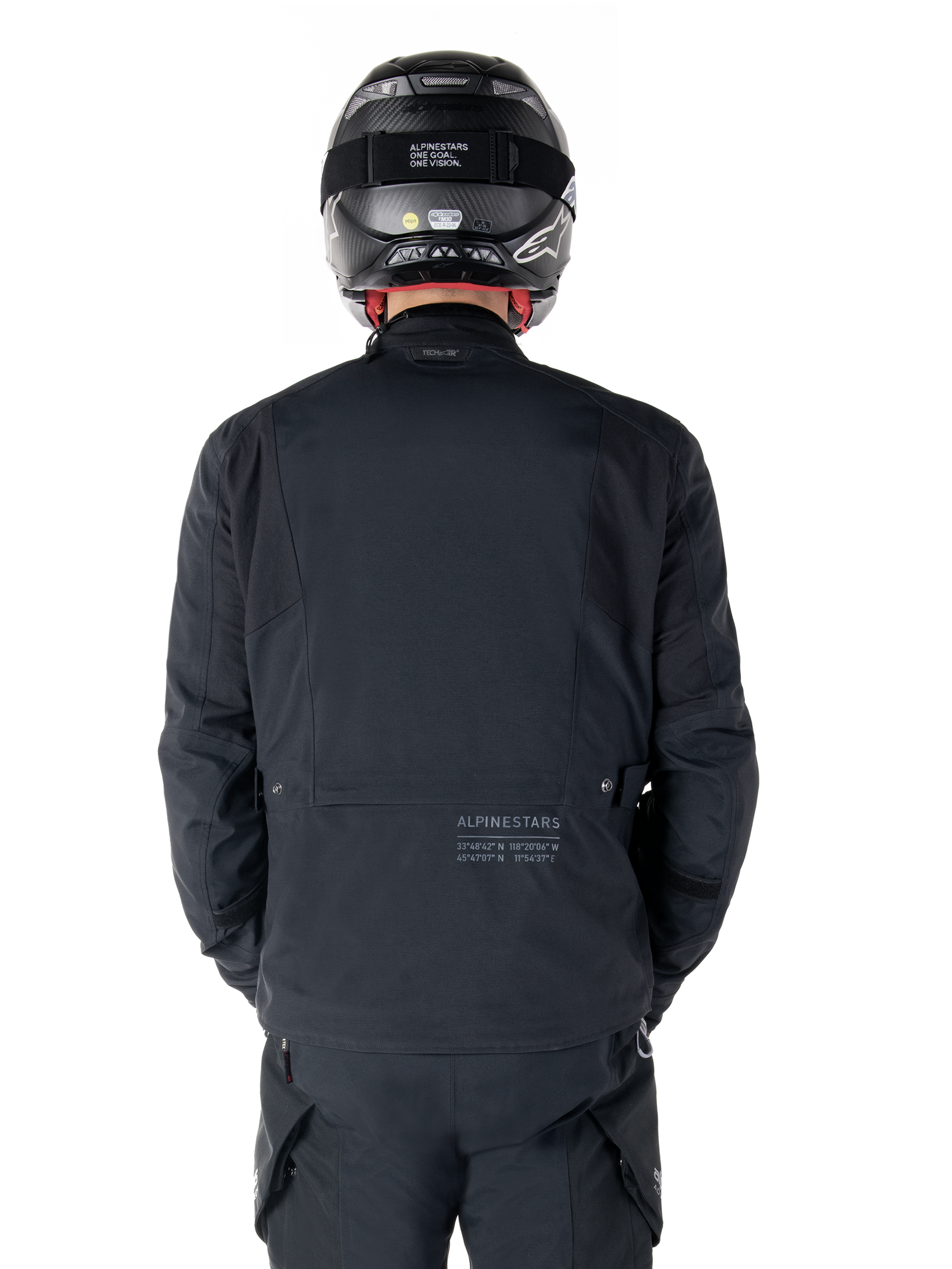 Alpinestars St-7 Prime GORE-TEX Jacket, Black, rear view of a rider, featuring subtle logo and geographic coordinates on the lower back, worn with a matching black helmet and pants, sleek and technical motorcycling apparel