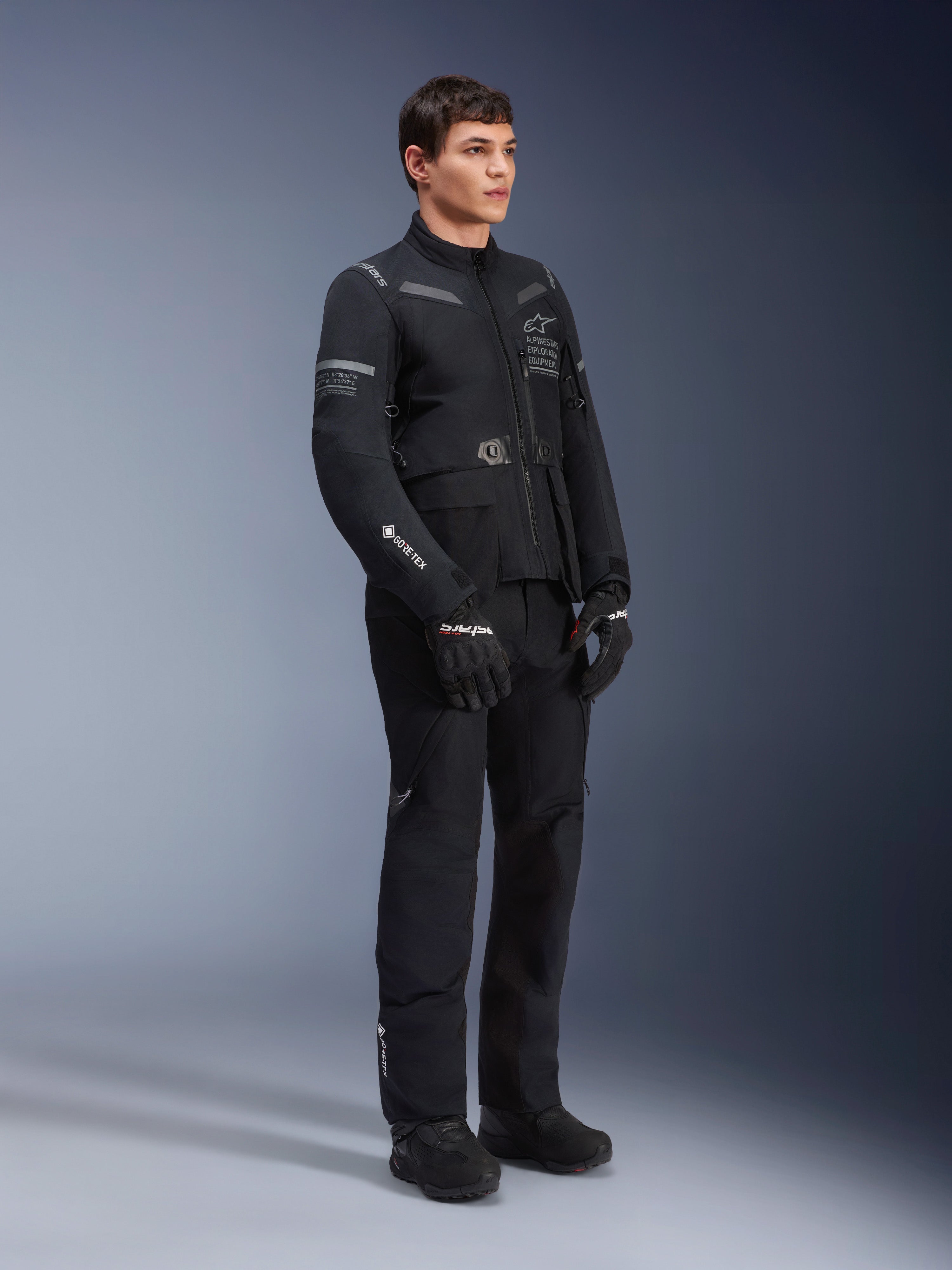 Modelo masculino con la chaqueta Alpinestars ST-7 2L GORE-TEX en color Black Dark Gray, chaqueta de moto para adventure touring, pantalones y guantes negros a juego, vista de tres cuartos de cuerpo completo, con el logotipo de GORE-TEX y la marca Alpinestars Exploration Equipment