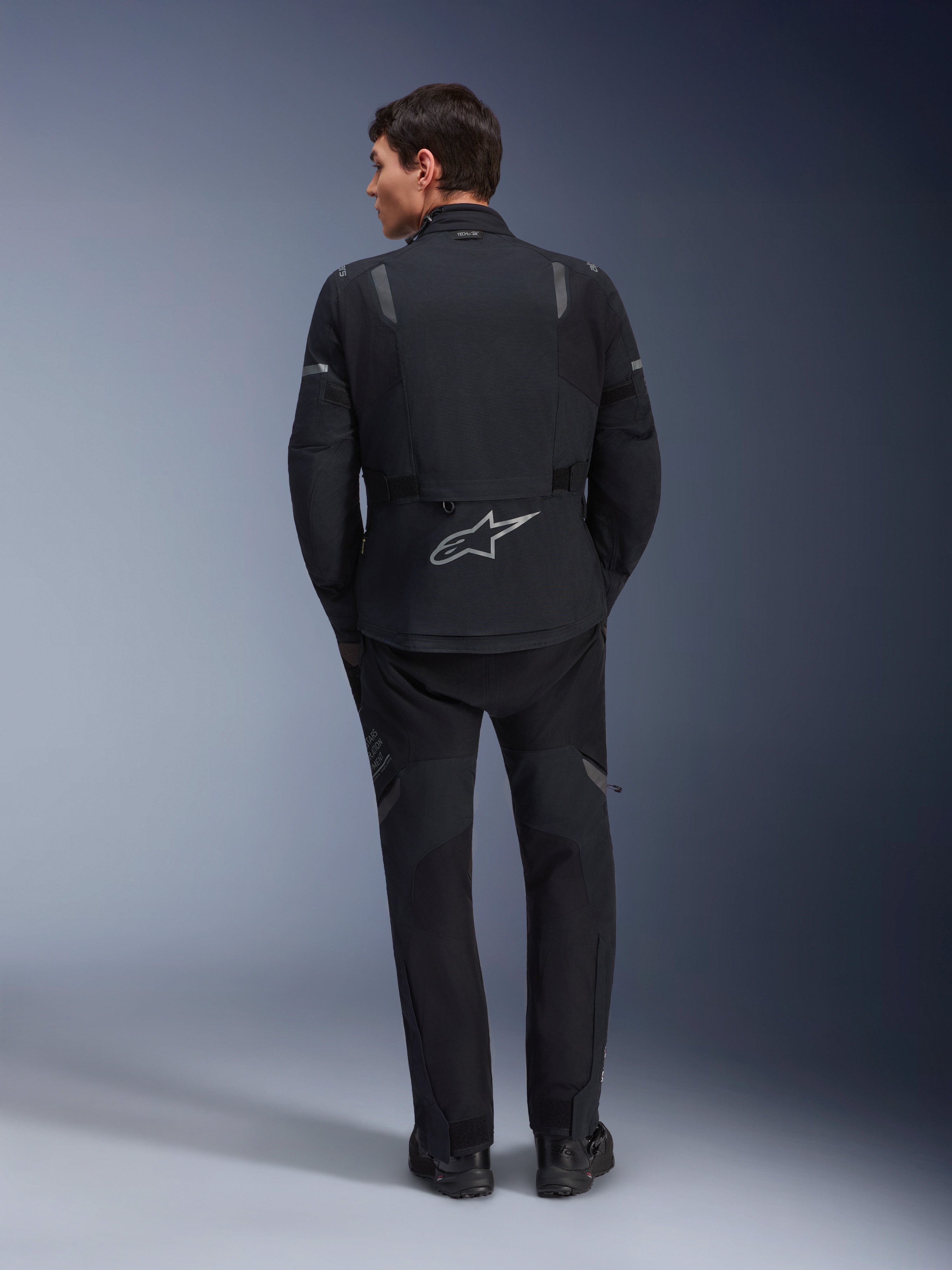 Chaqueta Alpinestars ST-7 2L GORE-TEX y pantalones ST-7 2L GORE-TEX a juego, equipamiento de moto de aventura negro y gris oscuro, vista trasera que muestra el logotipo reflectante de Alpinestars en la chaqueta y la construcción textil técnica.
