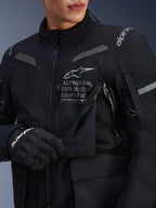 Primer plano de la chaqueta Alpinestars ST-7 2L GORE-TEX en negro y gris oscuro, con un panel de ventilación en el pecho abierto que muestra el forro de malla, el texto 'ALPINESTARS EXPLORATION EQUIPMENT' en el pecho y un guante Alpinestars negro.