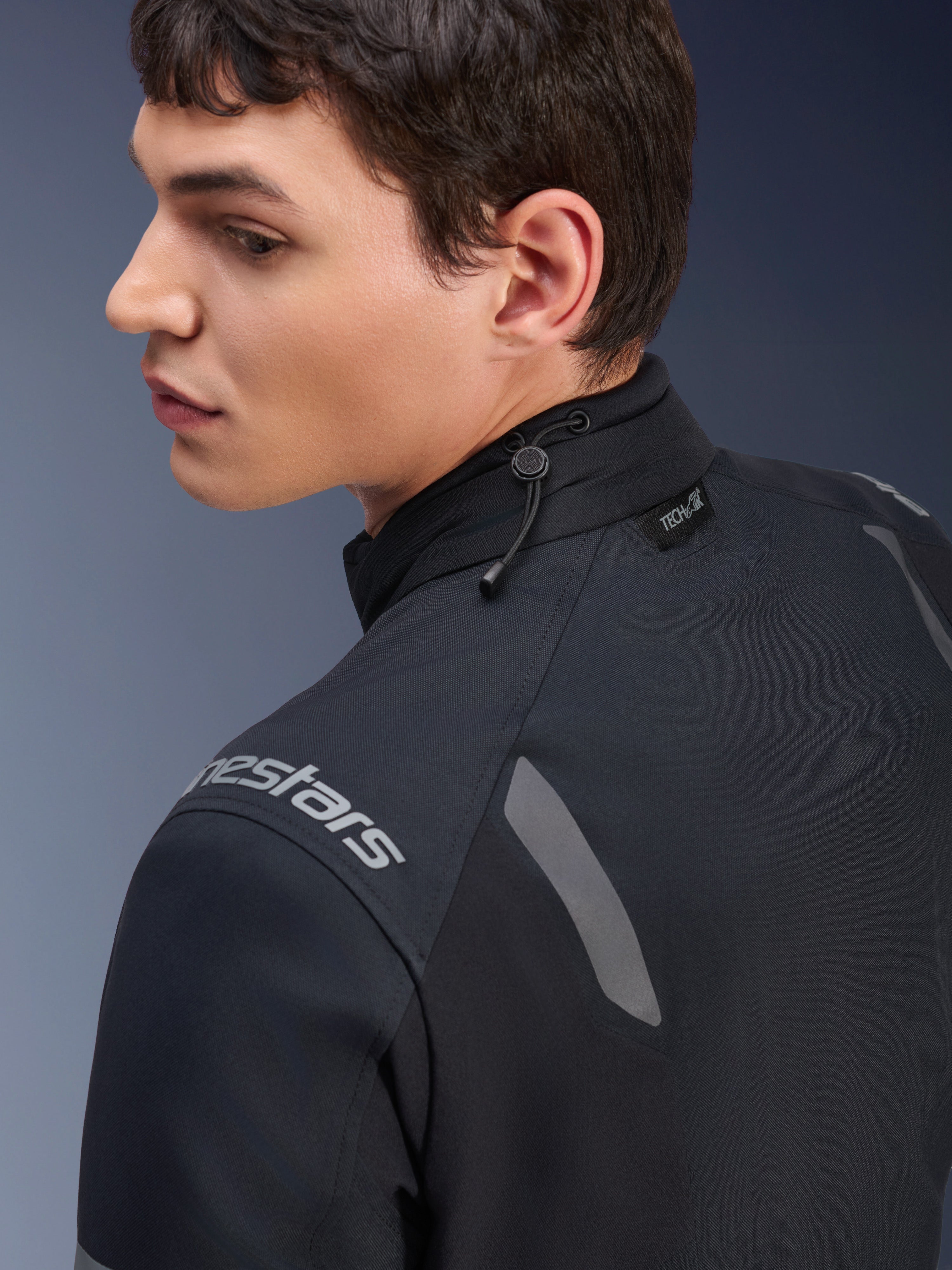 Chaqueta Alpinestars ST-7 2L GORE-TEX, chaqueta de motociclismo en negro y gris oscuro, vista de cerca del cuello que muestra el ajuste de cordón elástico y la etiqueta Tech-Air, con detalles reflectantes grises y logotipo de la marca en el hombro.