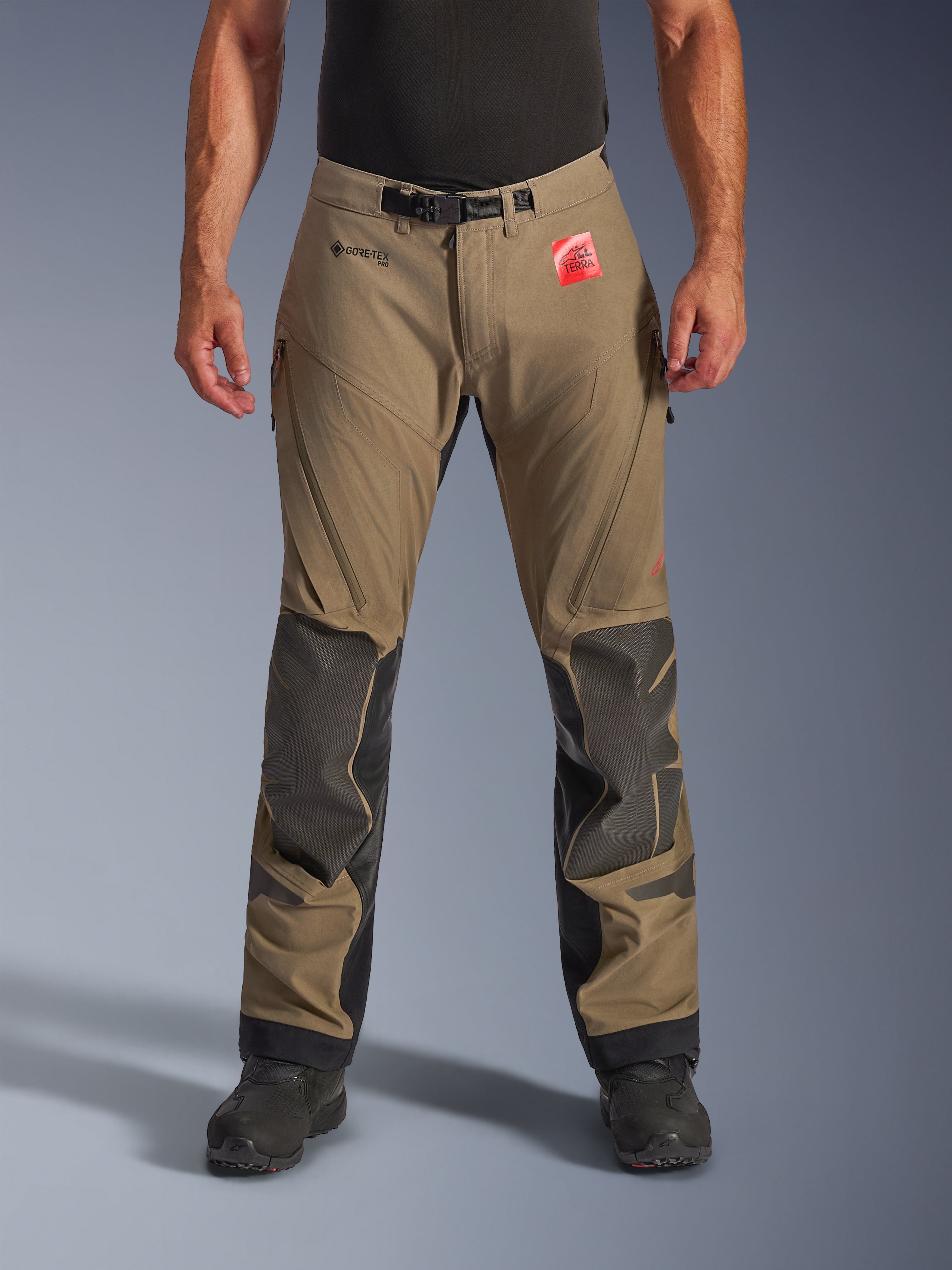 Alpinestars Nazca 3L GORE-TEX PRO Pants en Walnut Black, pantalones de Adventure Touring para hombre, con tejido marrón y refuerzos Superfabric negros en las rodillas, ventilaciones con cremallera en los muslos y cierre magnético en la cintura, vista frontal en modelo con botas negras, equipamiento de moto impermeable y duradero