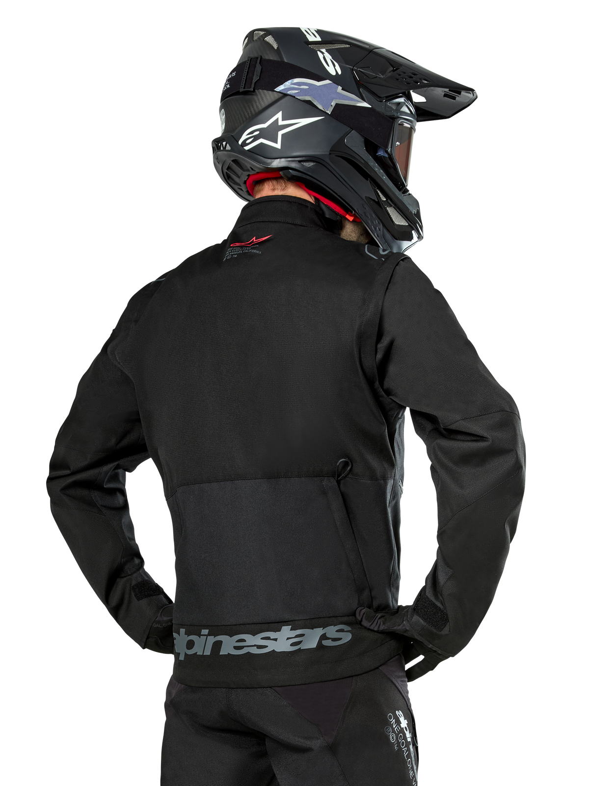 Vista posterior de un piloto con la chaqueta Alpinestars Pro-Dura en color negro, chaqueta de enduro de tejido ripstop duradero con un gran bolsillo de carga trasero y la marca Alpinestars en gris en la parte inferior de la espalda, combinada con un casco de motocross negro.