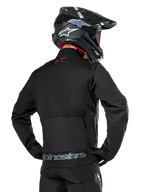 Vista posterior de un piloto con la chaqueta Alpinestars Pro-Dura en color negro, chaqueta de enduro de tejido ripstop duradero con un gran bolsillo de carga trasero y la marca Alpinestars en gris en la parte inferior de la espalda, combinada con un casco de motocross negro.
