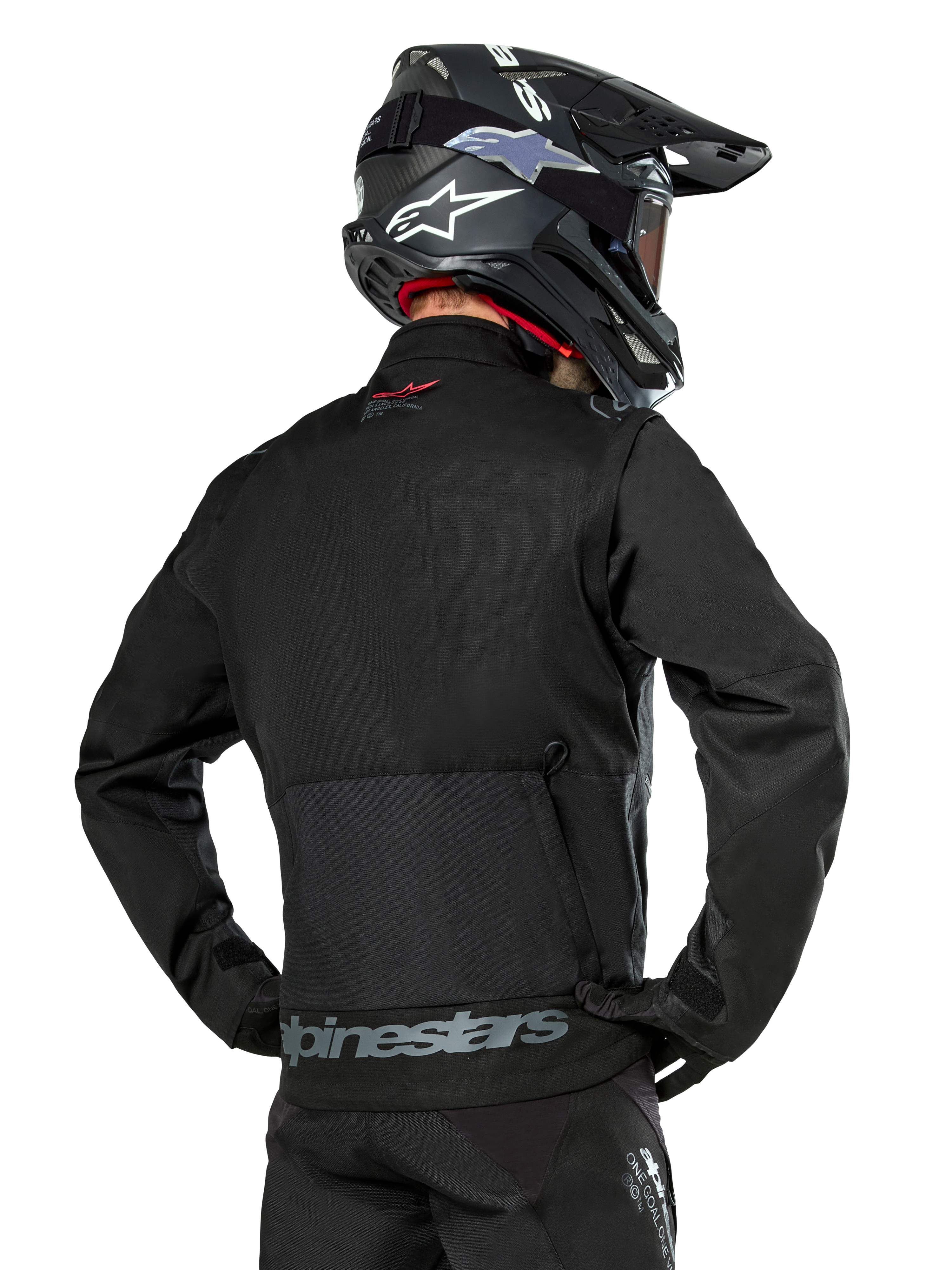 Vista posterior de un piloto con la chaqueta Alpinestars Pro-Dura en color negro, chaqueta de enduro de tejido ripstop duradero con un gran bolsillo de carga trasero y la marca Alpinestars en gris en la parte inferior de la espalda, combinada con un casco de motocross negro.