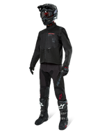 Piloto de motocross con equipamiento completo de carreras, chaqueta Alpinestars Pro-Dura en negro, pantalones negros con logotipos grises y detalles rojos, casco negro con gafas, y botas de motocross en blanco y negro, conjunto coordinado para enduro y conducción off-road