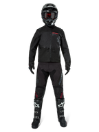 Chaqueta Alpinestars Pro-Dura, chaqueta de enduro off-road negra, mostrada en un modelo con un conjunto completo de equipamiento que incluye casco negro con gafas rojas, pantalones negros con detalles rojos, botas negras y blancas, y guantes negros, diseño robusto inspirado en el rally para recorridos por senderos