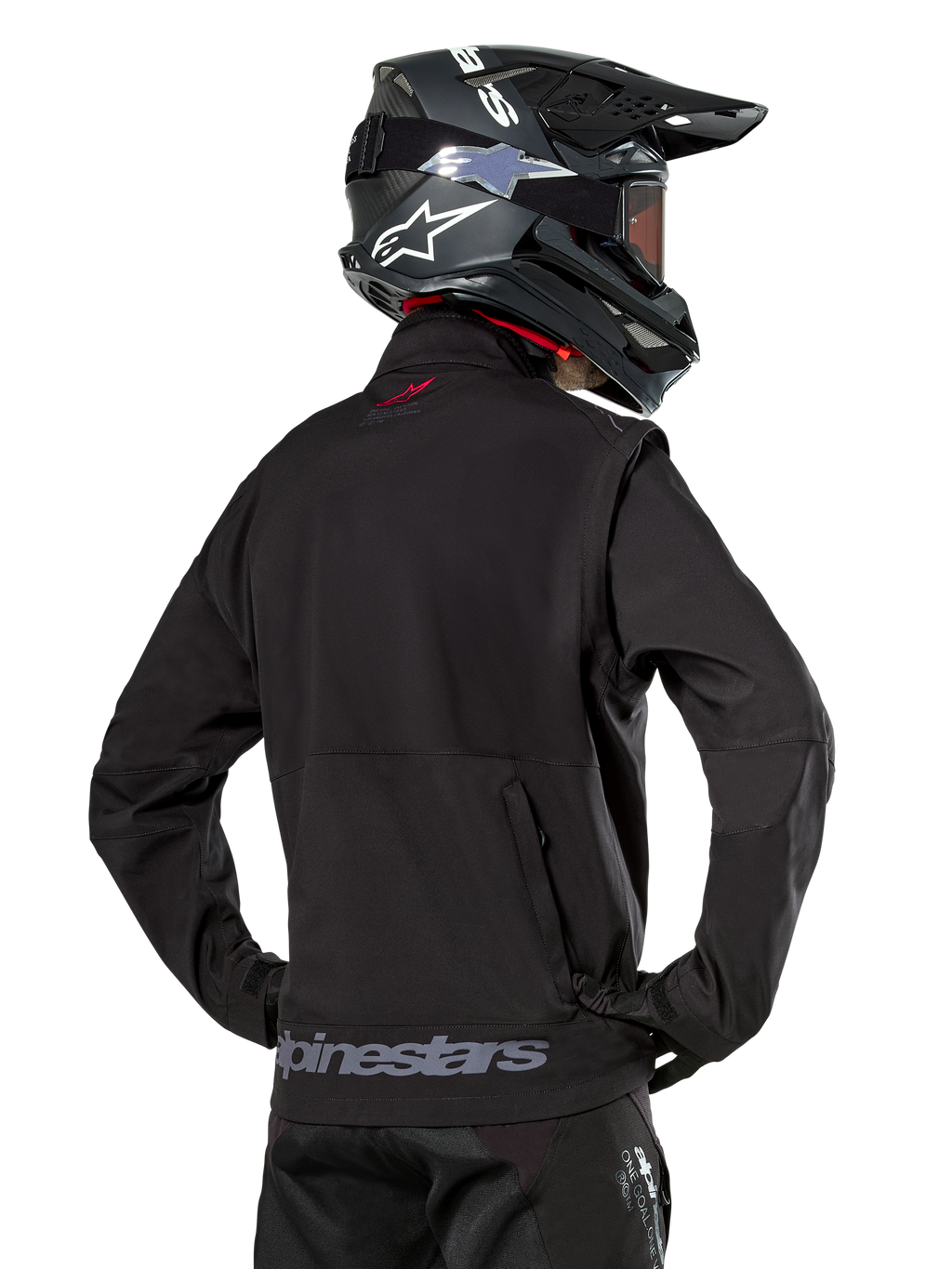 Chaqueta Softshell Alpinestars Lite-Dura, negra, vista trasera usada por un piloto, con marca Alpinestars gris en el dobladillo posterior y un pequeño logo rojo en el cuello, combinada con un casco de motocross negro, diseñada para enduro y conducción off-road