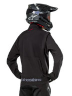 Chaqueta Softshell Alpinestars Lite-Dura, negra, vista trasera usada por un piloto, con marca Alpinestars gris en el dobladillo posterior y un pequeño logo rojo en el cuello, combinada con un casco de motocross negro, diseñada para enduro y conducción off-road