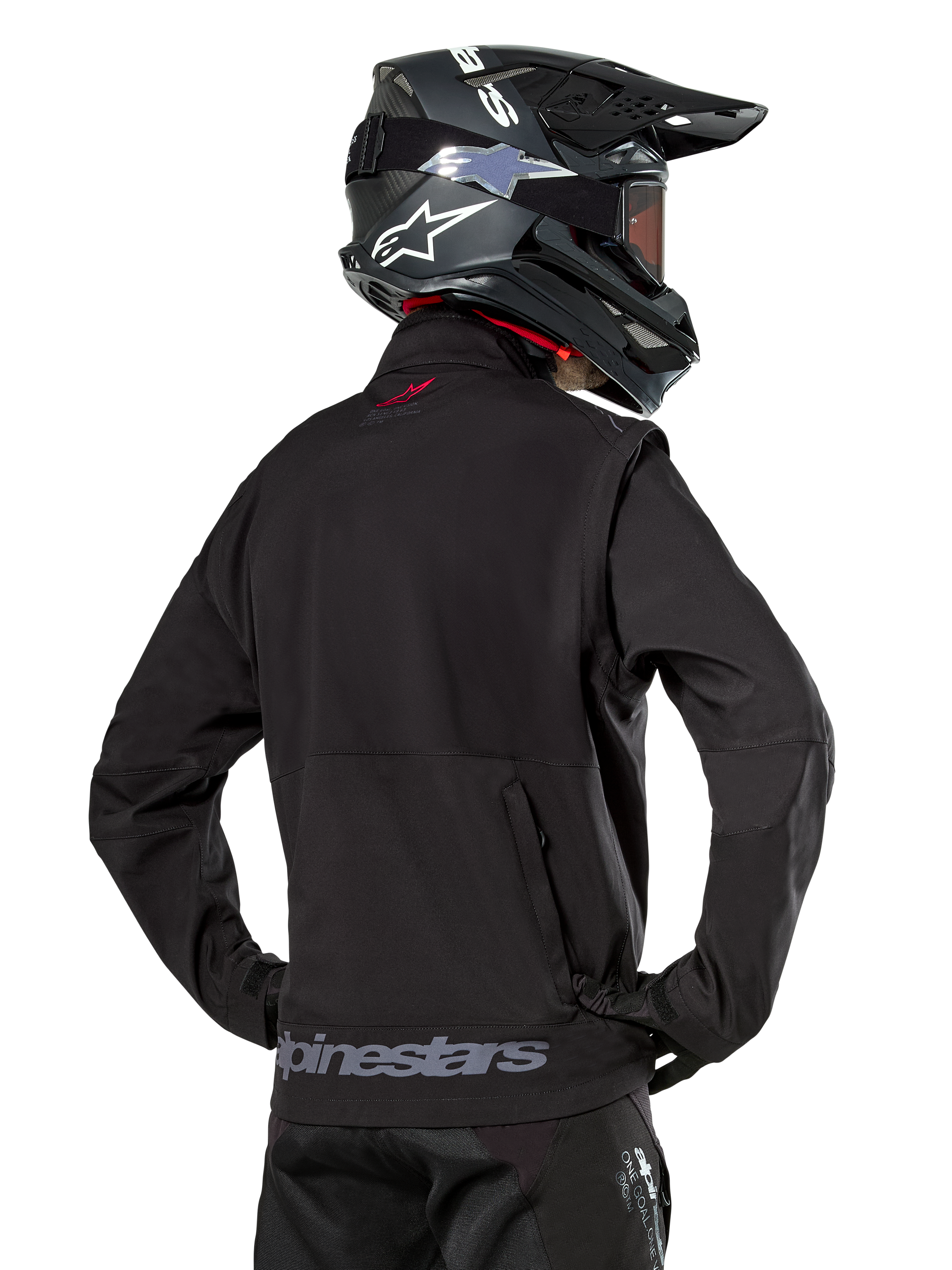Chaqueta Softshell Alpinestars Lite-Dura, negra, vista trasera usada por un piloto, con marca Alpinestars gris en el dobladillo posterior y un pequeño logo rojo en el cuello, combinada con un casco de motocross negro, diseñada para enduro y conducción off-road
