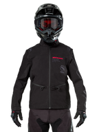 Piloto de motocross con la chaqueta Alpinestars Lite-Dura Softshell en color negro, detalles de logotipos en rojo y gris en el pecho, casco negro con gafas de lentes rojas, guantes y pantalones negros, vista frontal de equipamiento técnico para conducción off-road