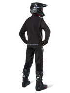 Piloto de enduro visto desde atrás con la chaqueta Alpinestars Lite-Dura Softshell en color negro, combinada con pantalones técnicos y botas negras, con un bolsillo de almacenamiento trasero con cremallera y el logotipo de Alpinestars en el dobladillo inferior, equipo completo de off-road para motocross.