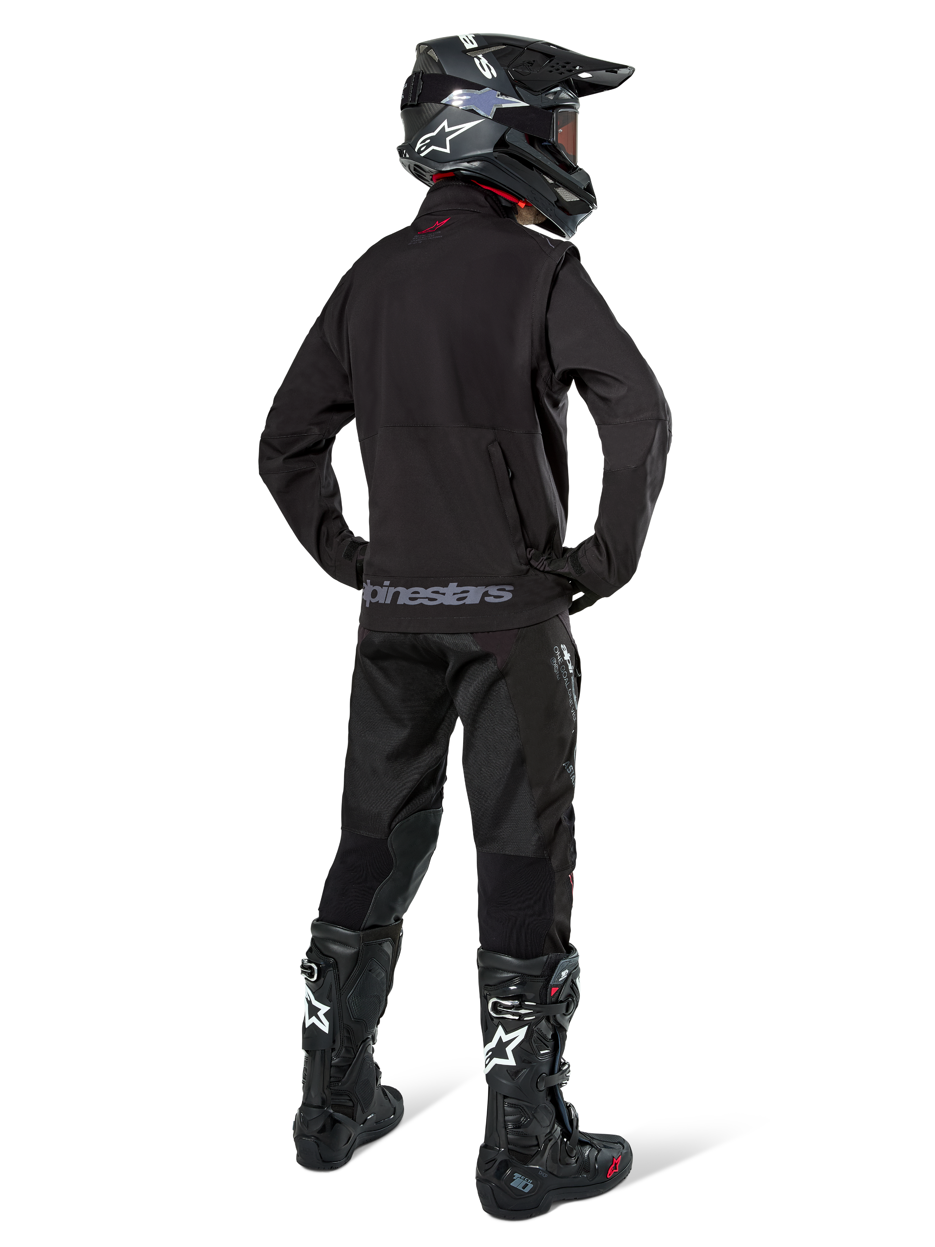 Piloto de enduro visto desde atrás con la chaqueta Alpinestars Lite-Dura Softshell en color negro, combinada con pantalones técnicos y botas negras, con un bolsillo de almacenamiento trasero con cremallera y el logotipo de Alpinestars en el dobladillo inferior, equipo completo de off-road para motocross.