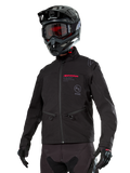 Alpinestars Lite-Dura Softshell Jacket, chaqueta de moto off-road negra con marca en rojo y logotipo AS, usada con casco de motocross negro, gafas rojas y guantes negros, con bolsillos con cremallera y puños ajustables, diseñada para enduro.