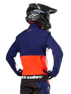 Chaqueta Softshell Alpinestars Lite-Dura, azul marino, naranja intenso y blanco, vista trasera de un piloto de motocross, con un gran logotipo blanco en el dobladillo inferior y bolsillo de almacenamiento con cremallera, parte de un equipo completo de carreras off-road.