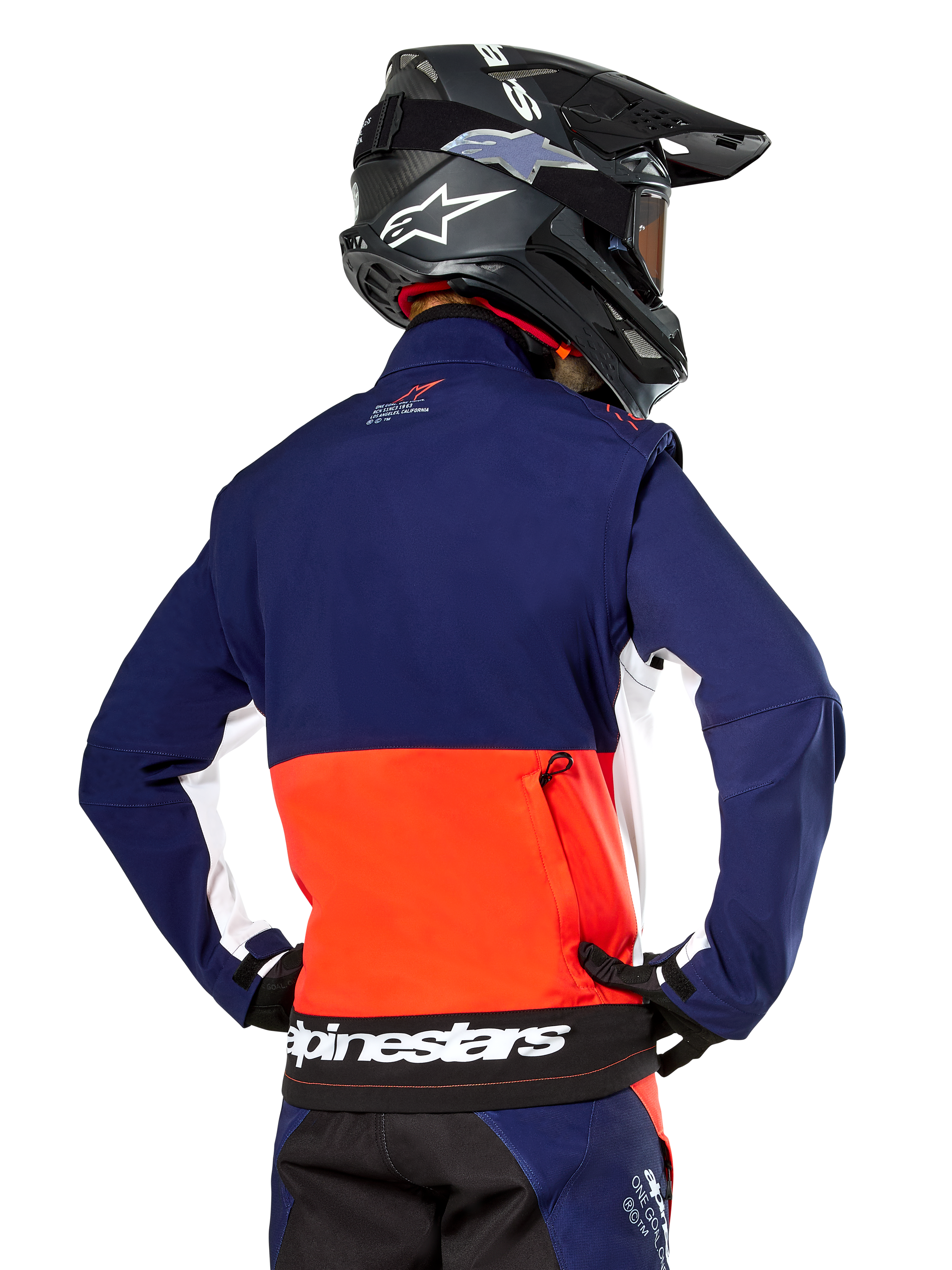 Chaqueta Softshell Alpinestars Lite-Dura, azul marino, naranja intenso y blanco, vista trasera de un piloto de motocross, con un gran logotipo blanco en el dobladillo inferior y bolsillo de almacenamiento con cremallera, parte de un equipo completo de carreras off-road.