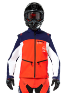 Chaqueta Softshell Alpinestars Lite-Dura en azul marino, naranja intenso y blanco, vista frontal de un piloto con casco negro y gafas, con diseño técnico de bloques de color, bolsillos con cremallera y logotipos de Alpinestars, equipamiento versátil para enduro y conducción off-road