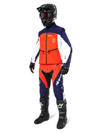 Piloto de motocross con la chaqueta Alpinestars Lite-Dura Softshell en azul marino, naranja intenso y blanco, pantalones de off-road a juego, casco negro con gafas y botas de carreras negras, conjunto completo de equipamiento de enduro