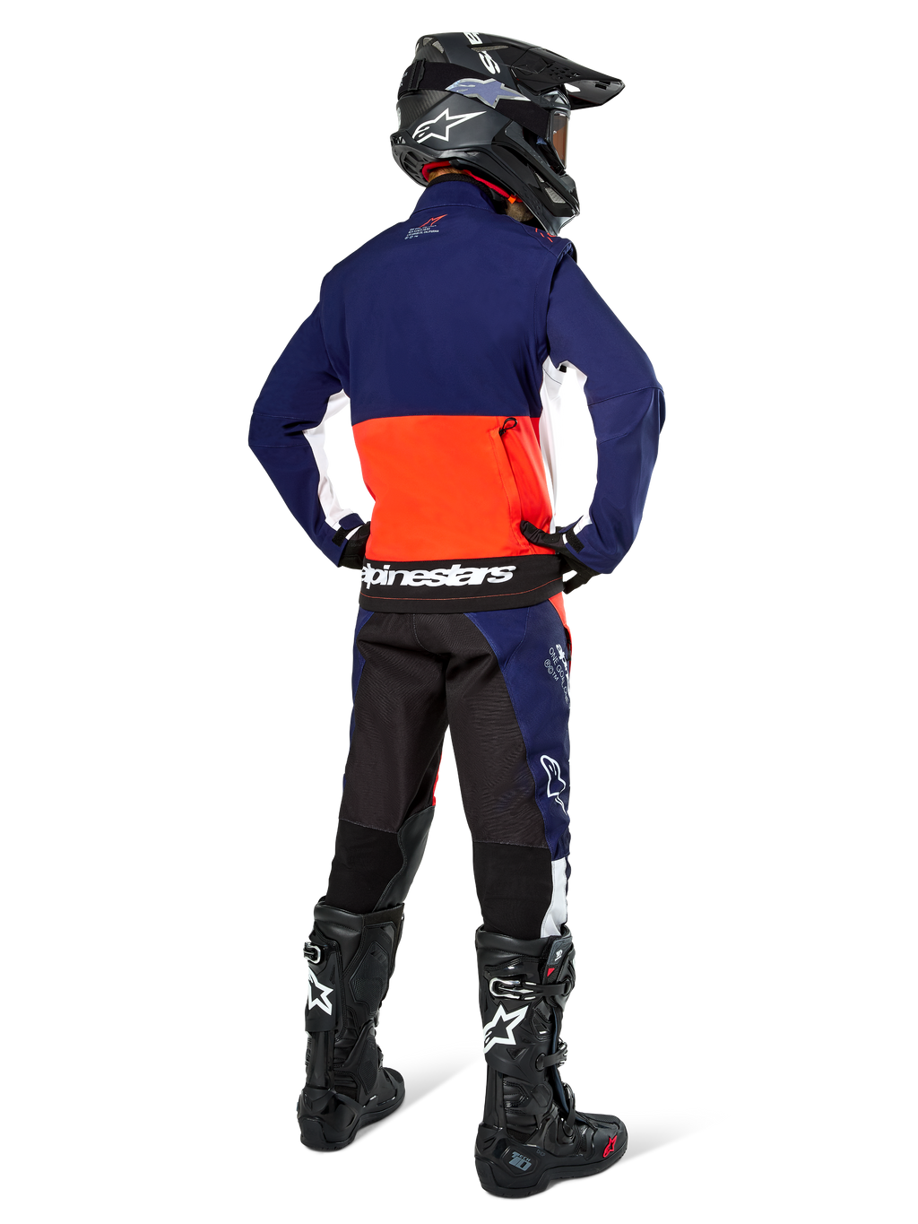 Piloto de motocross desde atrás vistiendo la chaqueta Alpinestars Lite-Dura Softshell en azul marino, naranja intenso y blanco, mostrando el gran bolsillo trasero; combinada con pantalones de enduro a juego, casco de motocross negro y botas off-road negras