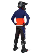 Piloto de motocross desde atrás vistiendo la chaqueta Alpinestars Lite-Dura Softshell en azul marino, naranja intenso y blanco, mostrando el gran bolsillo trasero; combinada con pantalones de enduro a juego, casco de motocross negro y botas off-road negras