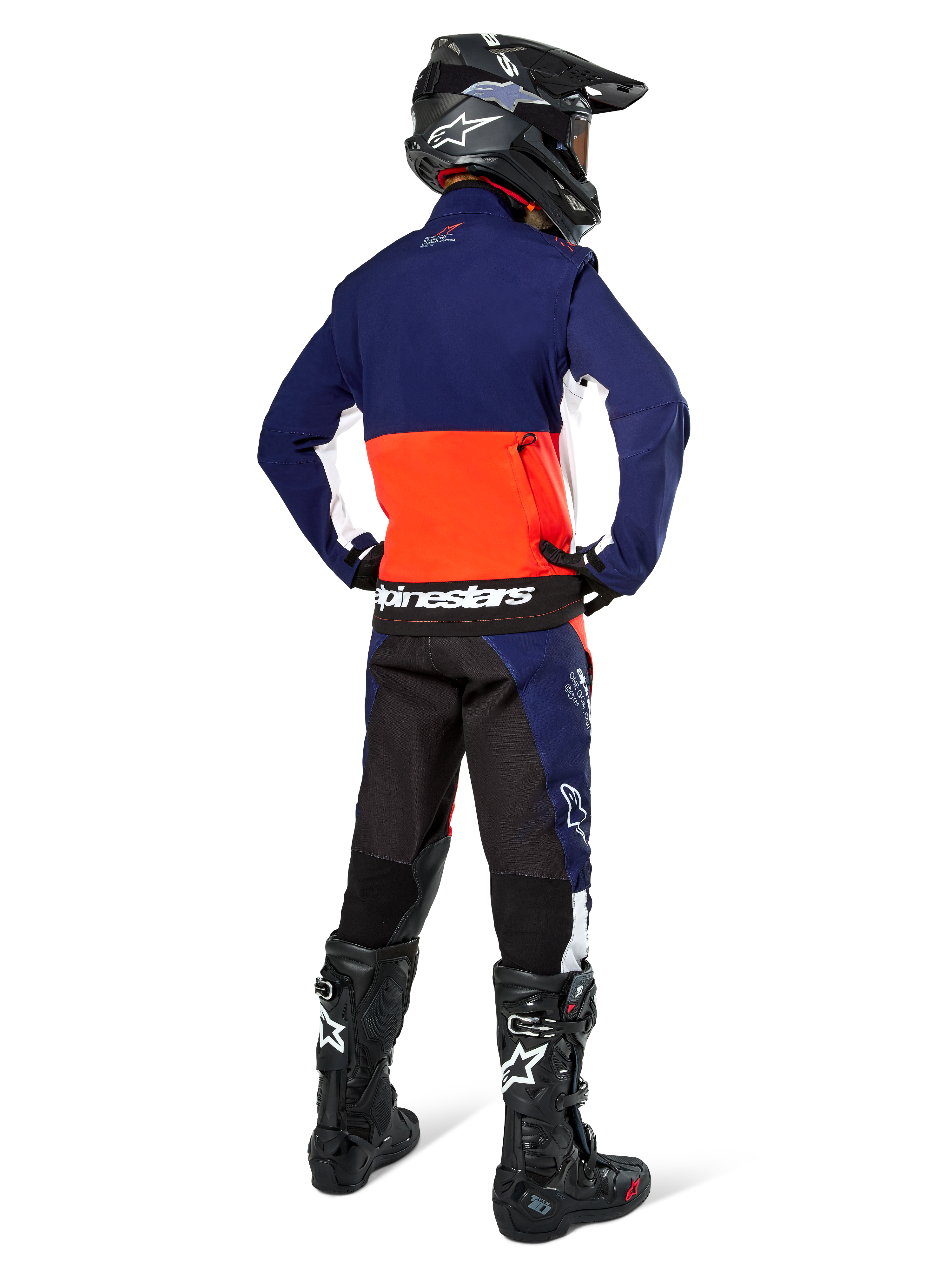 Piloto de motocross desde atrás vistiendo la chaqueta Alpinestars Lite-Dura Softshell en azul marino, naranja intenso y blanco, mostrando el gran bolsillo trasero; combinada con pantalones de enduro a juego, casco de motocross negro y botas off-road negras