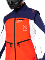 Chaqueta Softshell Alpinestars Lite-Dura en azul marino, naranja vibrante y blanco, vista en primer plano del torso, con un panel frontal naranja brillante, paneles laterales blancos y hombros azul marino, bolsillos para las manos con cremallera y marca Alpinestars destacada, usada por un piloto con un casco negro de off-road.