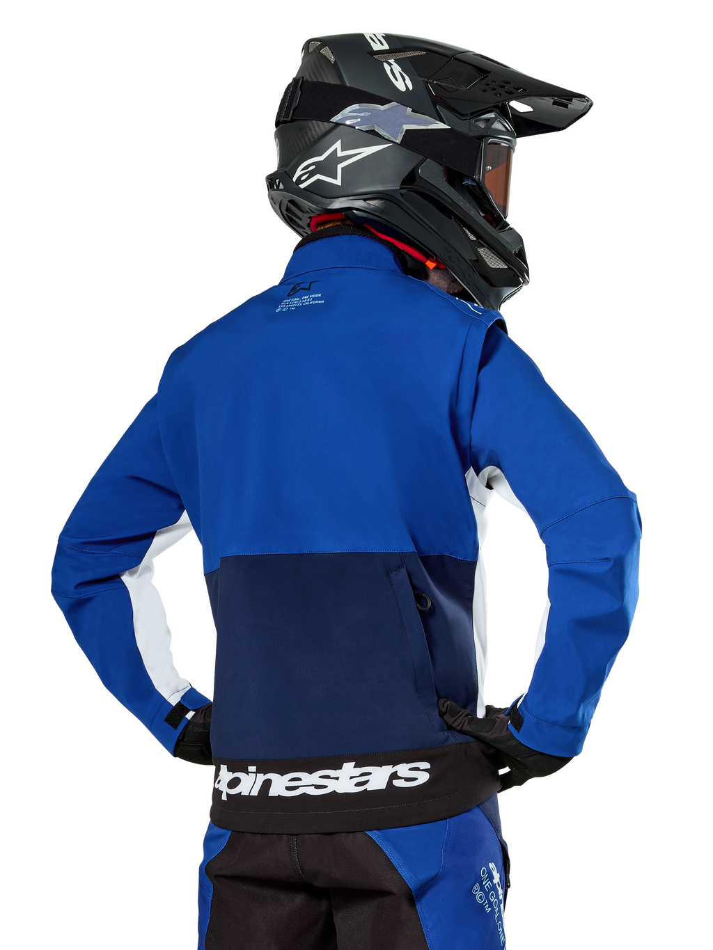 Chaqueta Softshell Alpinestars Lite-Dura en azul marino, azul brillante y blanco, vista trasera que muestra el gran bolsillo posterior con cremallera para guardar las mangas, detalles laterales blancos y marca Alpinestars en el dobladillo, usada con casco de motocross negro y equipamiento a juego