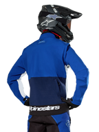 Chaqueta Softshell Alpinestars Lite-Dura en azul marino, azul brillante y blanco, vista trasera que muestra el gran bolsillo posterior con cremallera para guardar las mangas, detalles laterales blancos y marca Alpinestars en el dobladillo, usada con casco de motocross negro y equipamiento a juego
