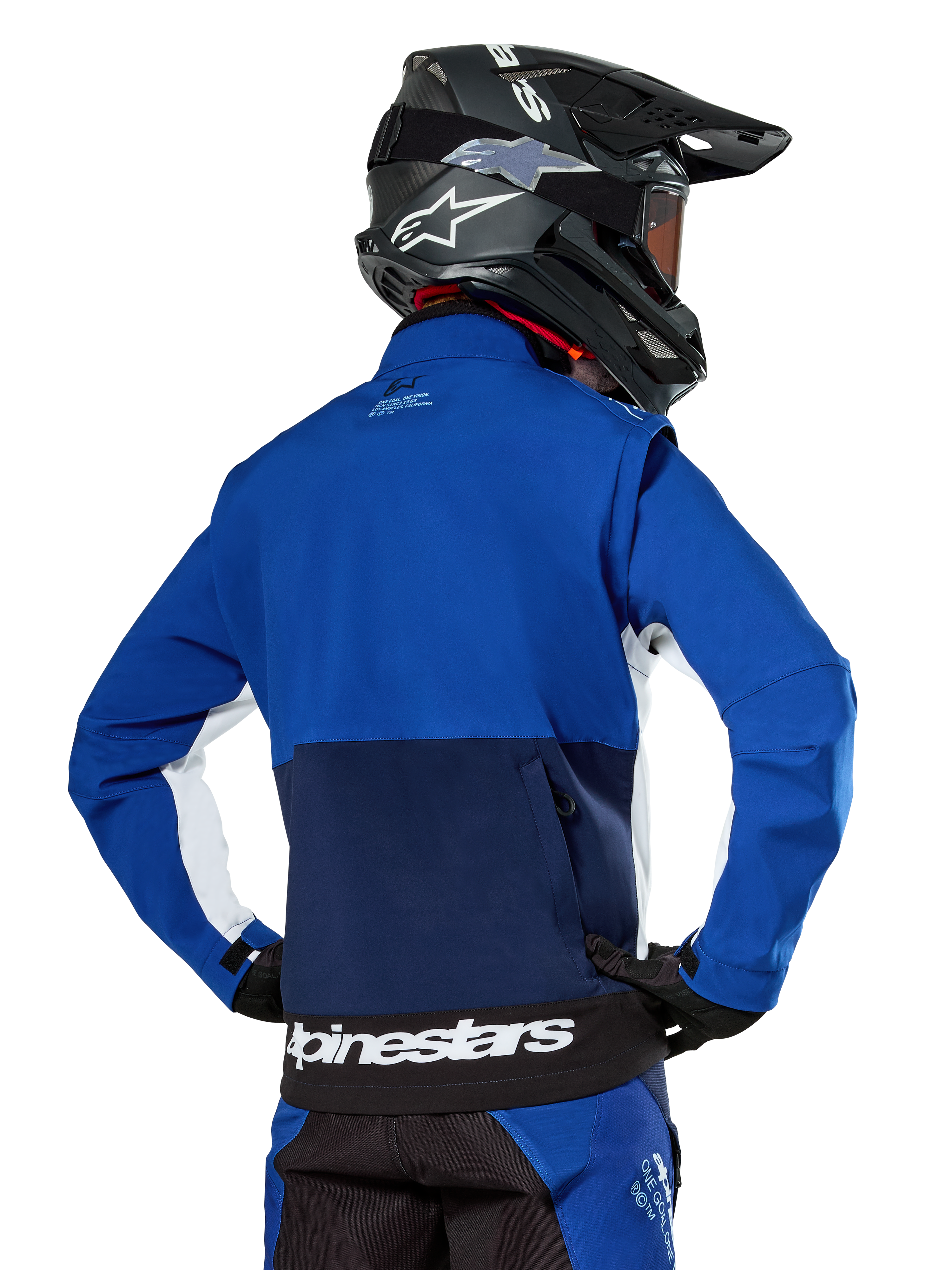 Chaqueta Softshell Alpinestars Lite-Dura en azul marino, azul brillante y blanco, vista trasera que muestra el gran bolsillo posterior con cremallera para guardar las mangas, detalles laterales blancos y marca Alpinestars en el dobladillo, usada con casco de motocross negro y equipamiento a juego