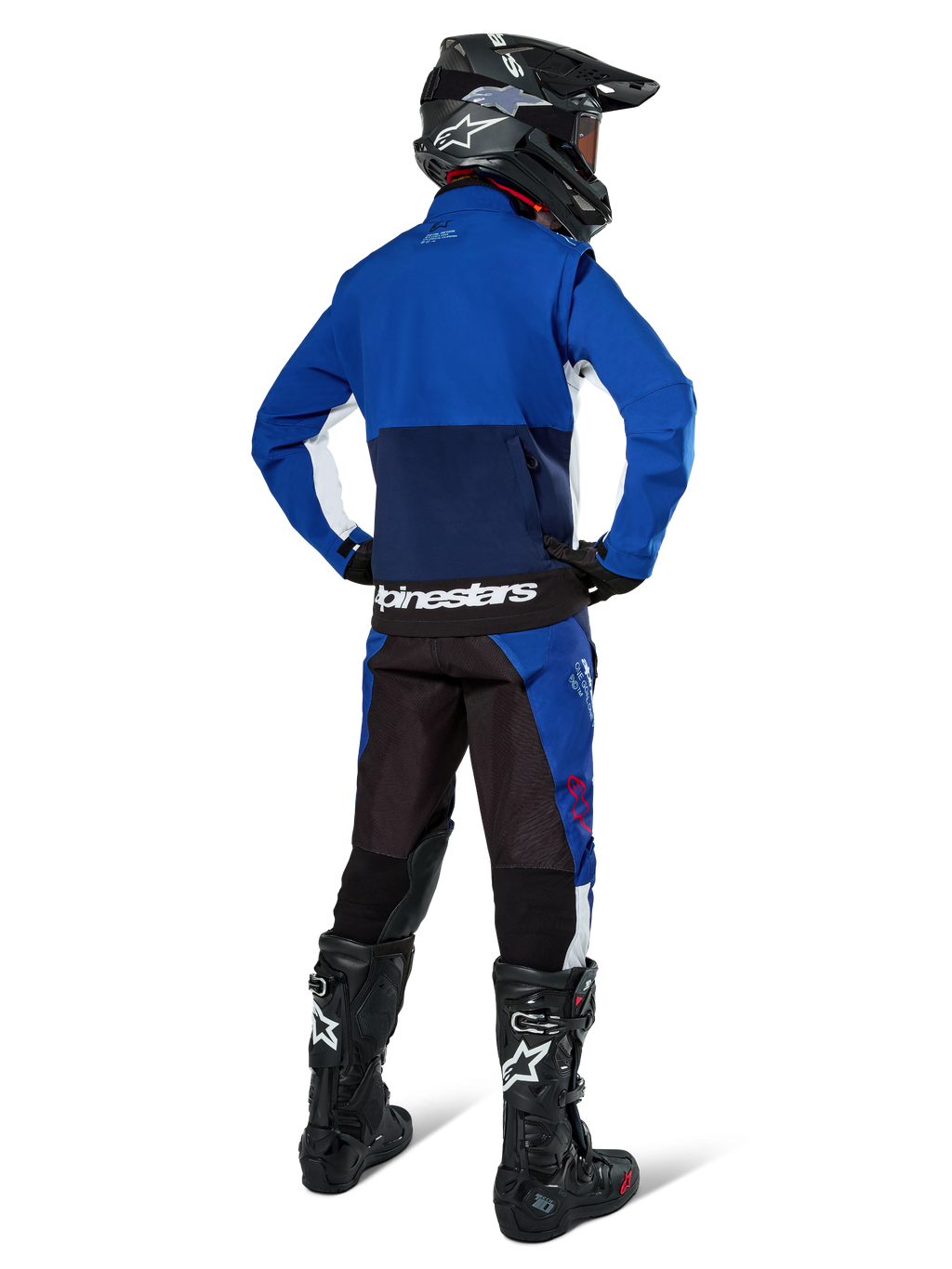 Piloto de motocross desde atrás vistiendo la chaqueta Alpinestars Lite-Dura Softshell en azul marino, azul brillante y blanco, combinada con pantalones de enduro a juego, casco negro y botas negras, equipo completo de carreras off-road
