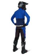Piloto de motocross desde atrás vistiendo la chaqueta Alpinestars Lite-Dura Softshell en azul marino, azul brillante y blanco, combinada con pantalones de enduro a juego, casco negro y botas negras, equipo completo de carreras off-road