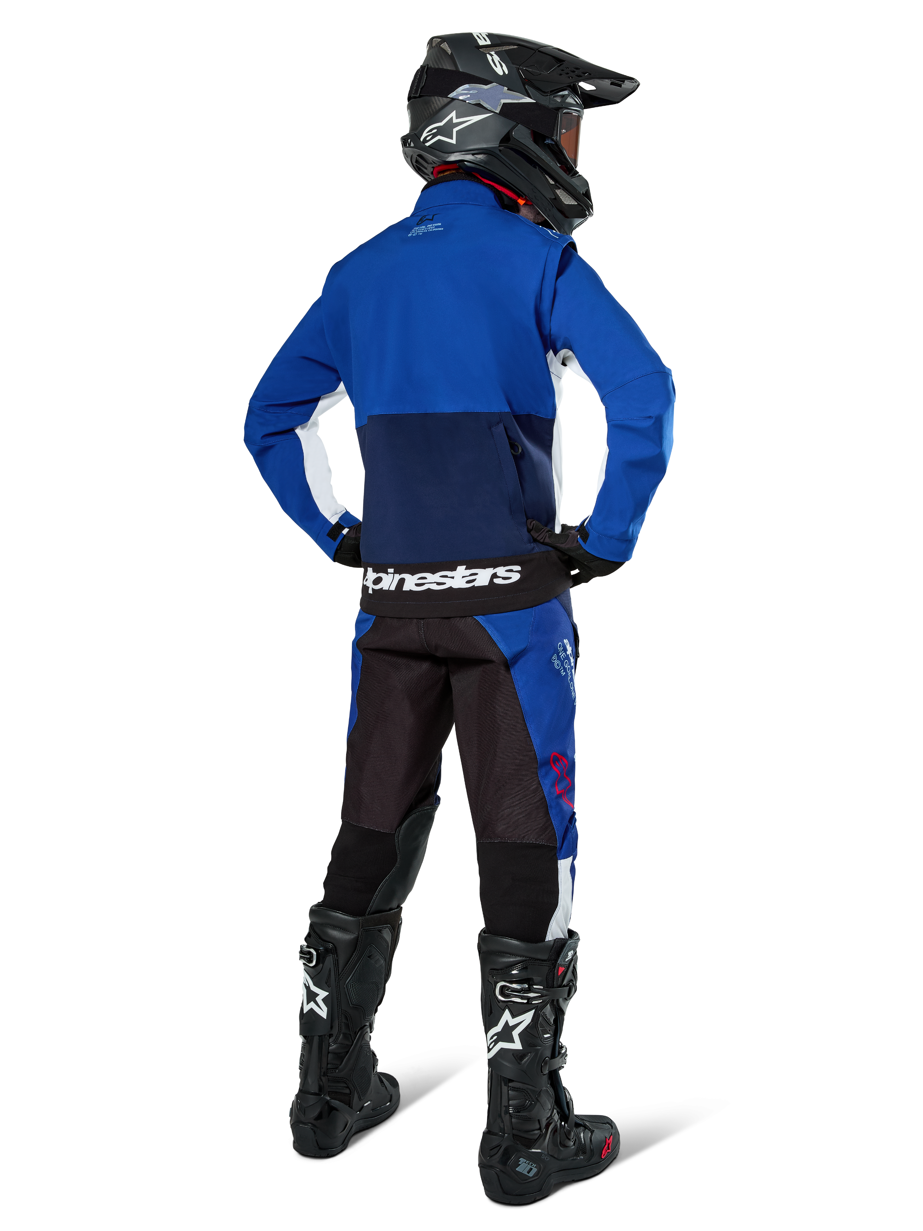 Piloto de motocross desde atrás vistiendo la chaqueta Alpinestars Lite-Dura Softshell en azul marino, azul brillante y blanco, combinada con pantalones de enduro a juego, casco negro y botas negras, equipo completo de carreras off-road