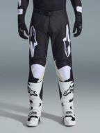 Alpinestars 2026 Racer Air Riway Pants, pantalones de motocross, negro y blanco, con llamativos logotipos blancos de Alpinestars en los muslos y paneles de las rodillas, combinados con botas de motocross blancas y negras y guantes negros, diseño atlético de ajuste de carrera para conducción off-road