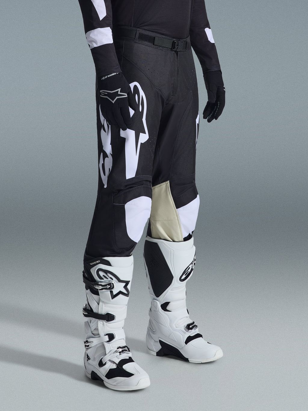 Alpinestars 2026 Racer Air Riway Pants, pantalones de motocross en blanco y negro con grandes gráficos del logotipo en blanco, paneles interiores de las rodillas en cuero beige, usados con botas de off-road blancas y negras y guantes negros en un entorno de estudio.