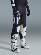 Alpinestars 2026 Racer Air Riway Pants, pantalones de motocross en blanco y negro con grandes gráficos del logotipo en blanco, paneles interiores de las rodillas en cuero beige, usados con botas de off-road blancas y negras y guantes negros en un entorno de estudio.