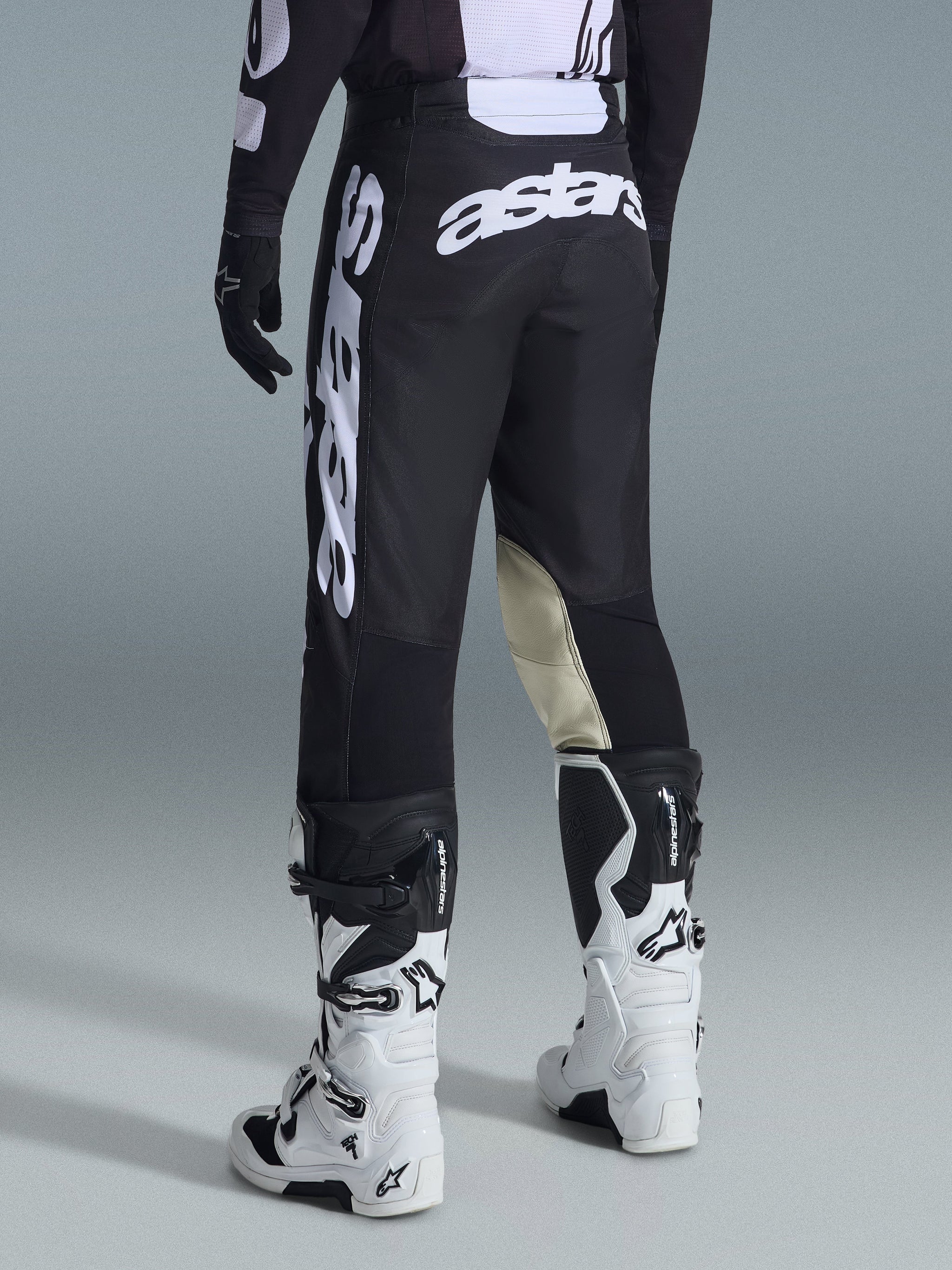 Alpinestars 2026 Racer Air Riway Pants, pantalones de motocross, negro y blanco, vista trasera con gran logotipo Astars blanco en el asiento y las piernas, paneles de cuero canela en la parte interna de las rodillas, combinados con botas Tech 7 blancas y negras, equipamiento premium para carreras todoterreno.