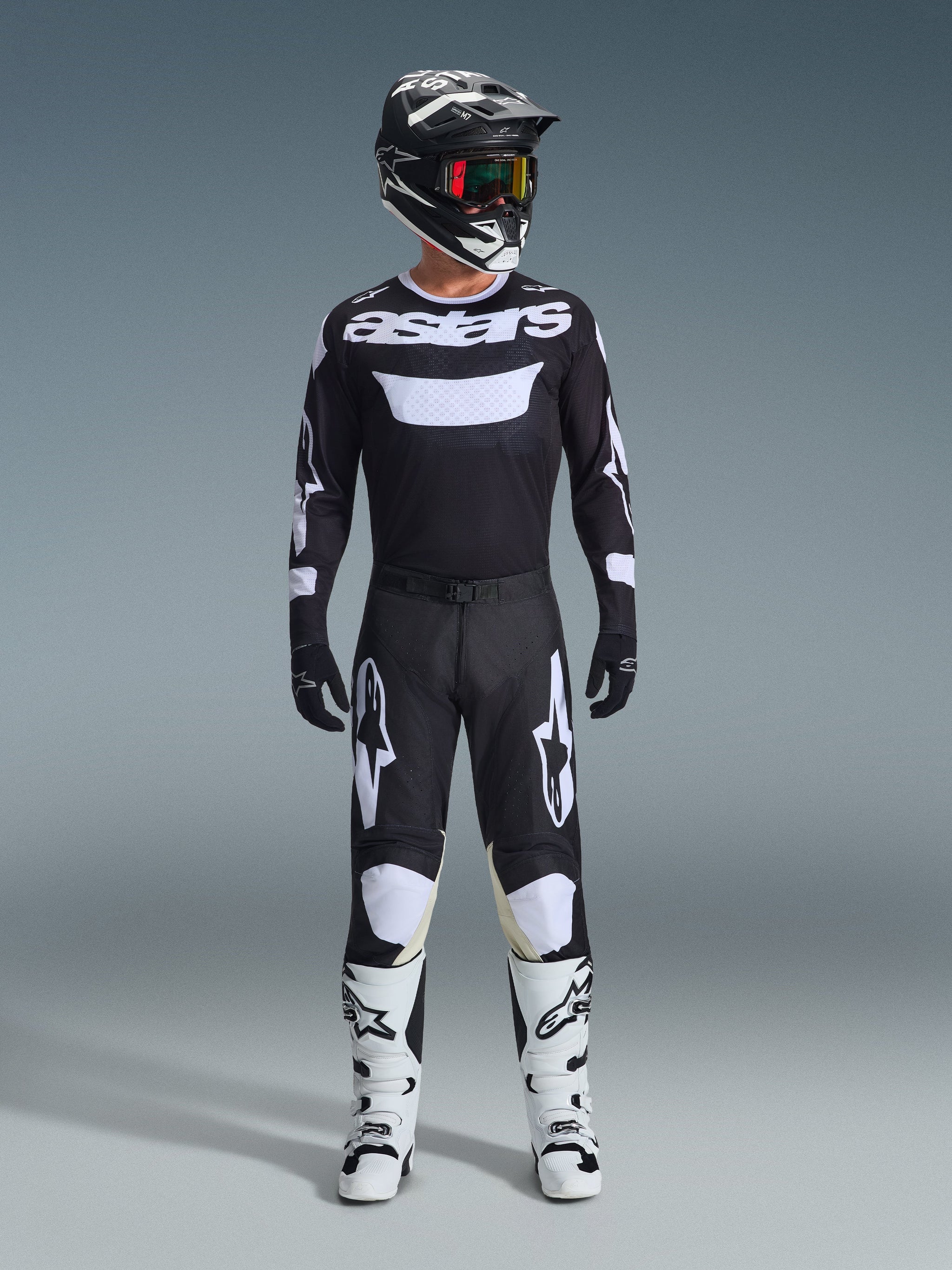 Piloto de motocross con pantalones Alpinestars Racer Air Riway 2026 en negro y blanco, jersey a juego con el logo 'astars', casco negro, gafas con lentes iridiscentes y botas de carreras blancas, conjunto completo de equipamiento profesional para off-road