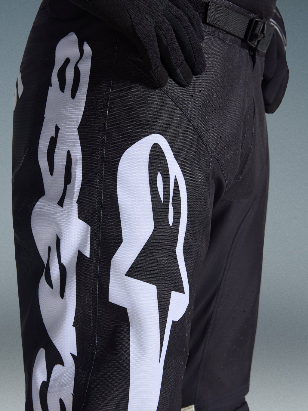 Alpinestars 2026 Racer Air Riway Pants, pantalones de motocross negros con un llamativo logotipo blanco de Alpinestars en el muslo, primer plano que muestra el tejido técnico y la correa ajustable de la cintura, usados con guantes negros para conducción off-road